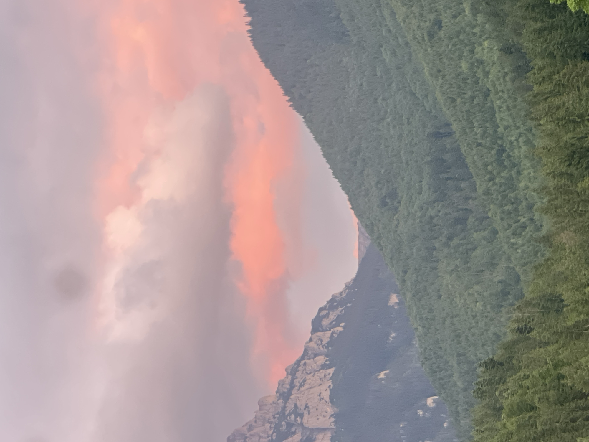 A mountain range is depicted at dusk. The sky is filled with clouds that are illuminated by the setting sun, casting hues of pink and orange. The mountains are covered in dense evergreen forests. On the left, a rocky mountain face is visible, contrasting with the softer green slopes. The scene is tranquil and natural, with no signs of human presence or urban development. The location is not identifiable as being within a specific city.