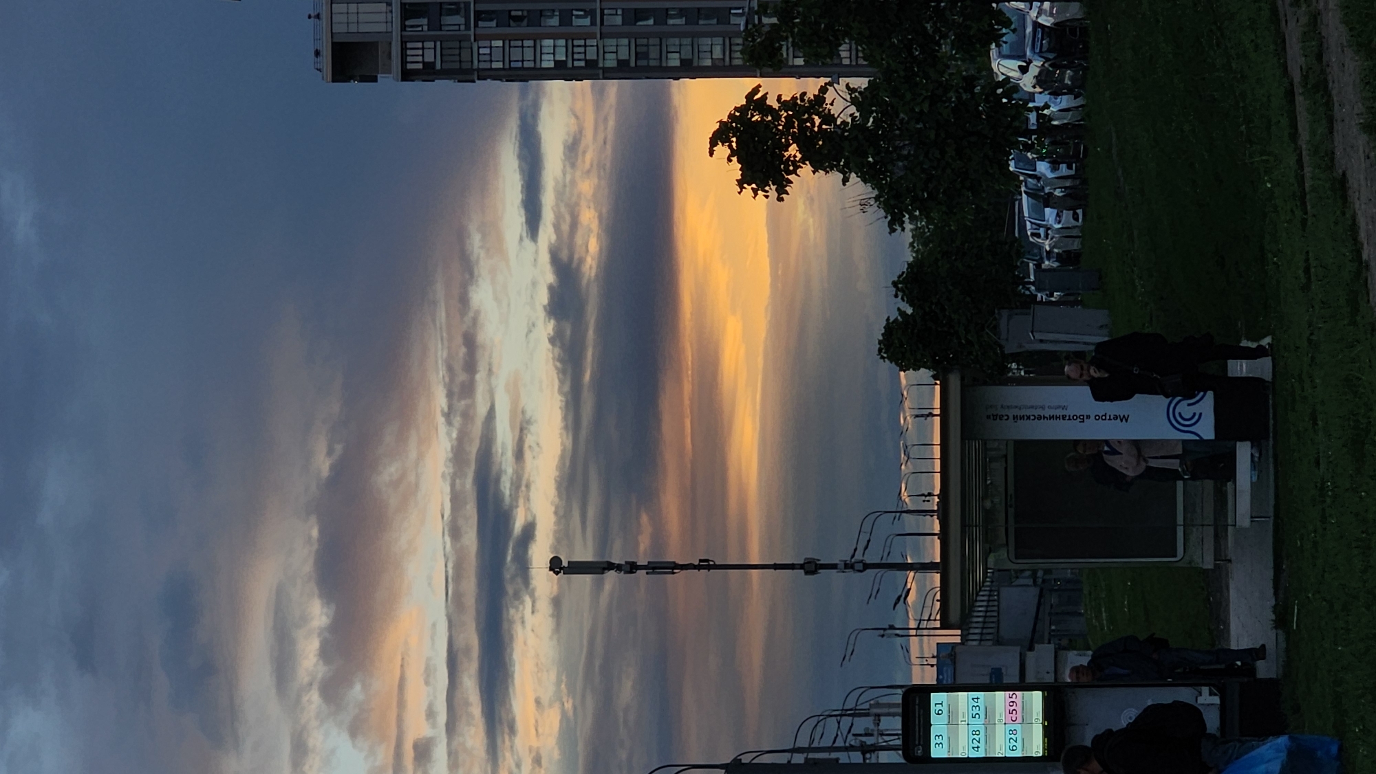 The image shows a street scene in Moscow, Russia, at dusk or dawn. The sky is filled with clouds streaked with the colors of the setting or rising sun. A tall building is visible in the upper left. On the right side of the image, there's a line of parked cars, grass, and a bus stop. People are standing and sitting near the bus stop. A sign at the bus stop reads "Metro 'Botanichesky Sad'", indicating the location is near the Botanichesky Sad (Botanical Garden) metro station. Streetlights line the road, and a tall communications tower is visible. A digital display is visible and shows bus numbers.