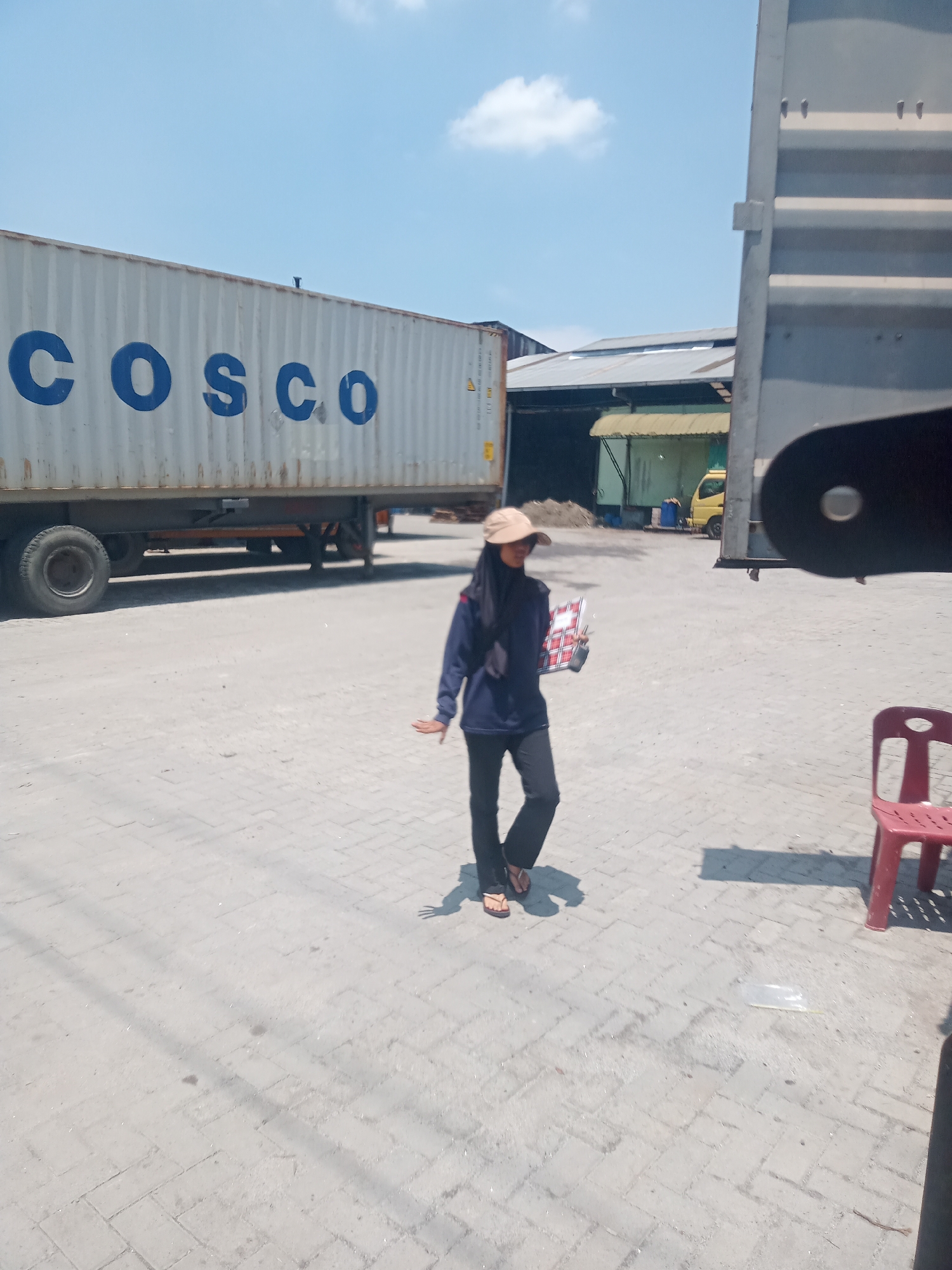 A person wearing a dark blue jacket, black pants, a black hijab, and a tan baseball cap walks in an outdoor industrial area. The person is holding a red and white patterned item. A white shipping container with "COSCO" written in blue is parked behind the person. A large, dark gray metal structure, possibly part of another container or vehicle, is to the right. The ground is paved with light gray interlocking bricks. In the background, there are buildings with gray roofs and a yellow truck. A red plastic chair is visible to the right of the person. The sky is blue with scattered white clouds. The scene appears to be in an industrial area in the city of Sunggal, Indonesia.