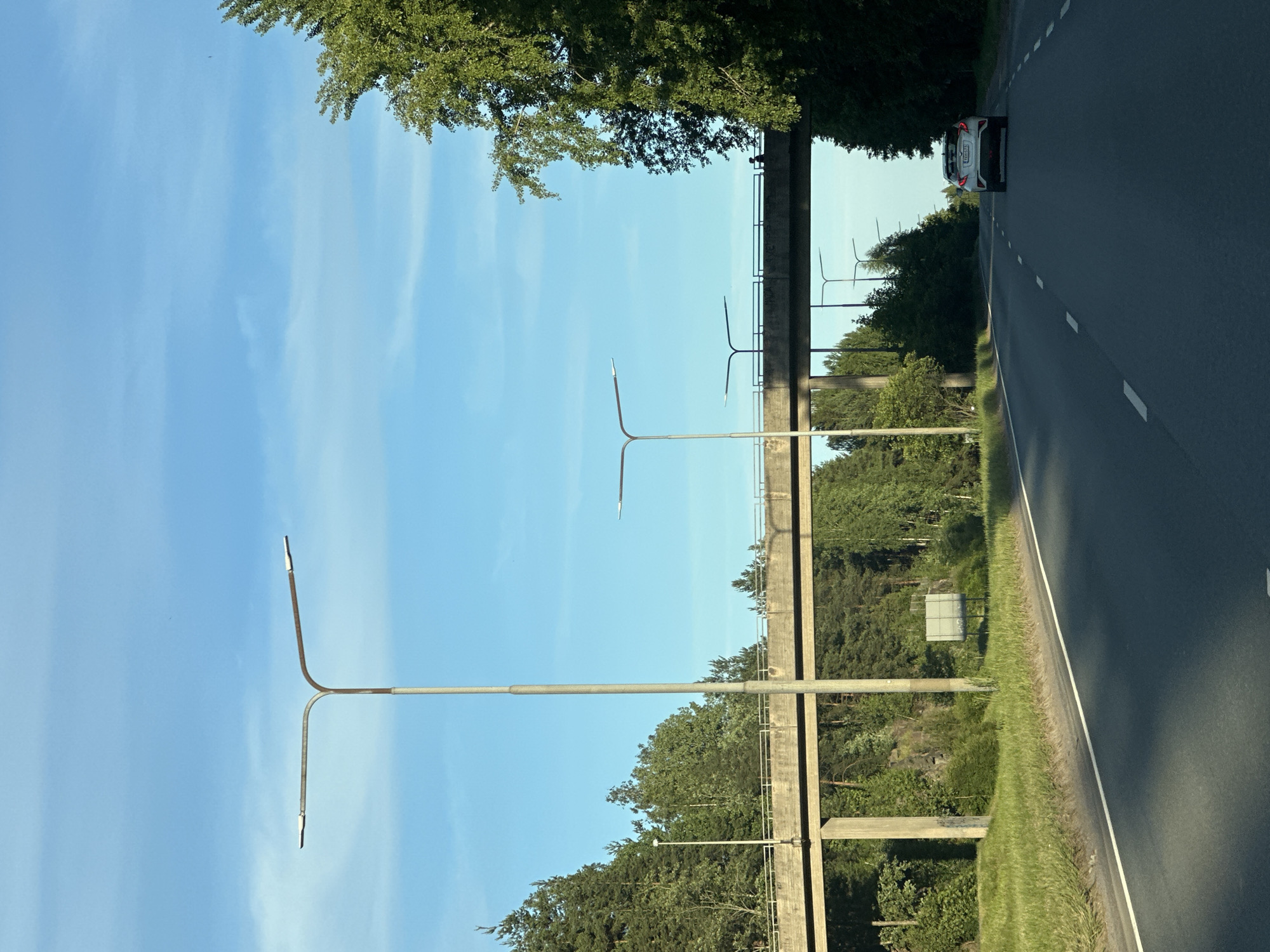 This media file depicts a roadside scene with a clear blue sky. The perspective is from a moving vehicle, looking towards the right side of the road. A white car is visible in the distance on the road, which has white dashed lines marking the lanes. On the left side of the road, there is an elevated concrete structure, possibly a bridge or overpass, with a railing. Several tall lampposts with a Y-shaped design are positioned along the road and under the overpass. Lush green trees and foliage line the roadside and surround the elevated structure. The lighting suggests it is daytime, with the sun likely casting soft shadows. The overall atmosphere is serene and suggests a typical day on a highway. No people are clearly visible, and no text is discernible within the image.