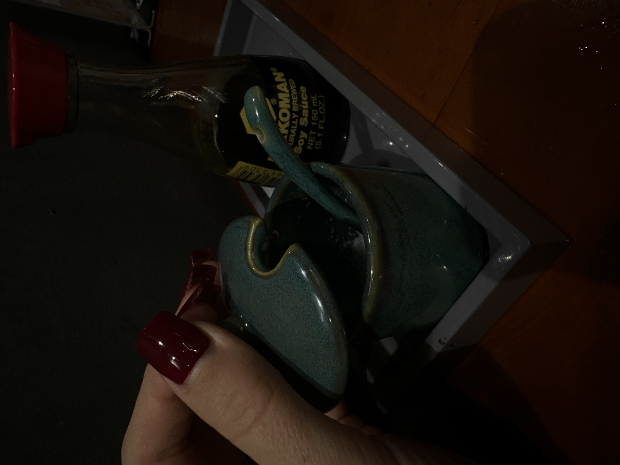 The image, captured in what is likely a dining setting in Nha Trang Ward, Vietnam, presents a close-up view of a person's hand interacting with condiments. The hand, adorned with long, dark red polished fingernails, is holding a distinctive teal or dark green ceramic container with a speckled, glossy finish.
In the background, a small glass bottle of Kikkoman Naturally Brewed Soy Sauce is visible, featuring the text "KIKKOMAN®", "NATURALLY BREWED", "Soy Sauce", and "NET 150 mL (5.1 FL. OZ.)". This bottle, along with another matching teal ceramic container (possibly a lidded pot with a small ceramic spoon), rests within a gray tray.
The overall scene is dimly lit, suggesting an evening setting or a restaurant with low artificial lighting. A dark surface forms the immediate background, while a warmer brown or wooden surface is partially visible further back. No specific weather conditions are discernible, and no emotions are overtly displayed.