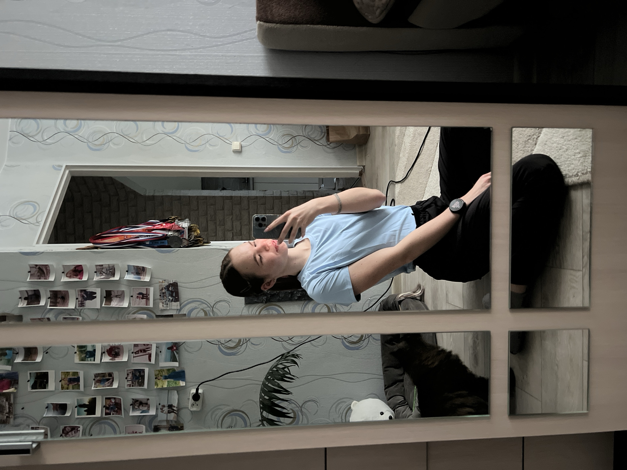 The image is a mirror selfie of a young woman in a room with a patterned wallpaper. She is wearing a light blue T-shirt and black pants, and she is holding a smartphone. A collection of photos, along with medals and keys, are pinned on the wall above her. The room's interior design includes a decorative leaf and a white bear figure. The reflection shows elements of the surrounding environment, including flooring and a rug. The location is Chaikovsky, Russia.