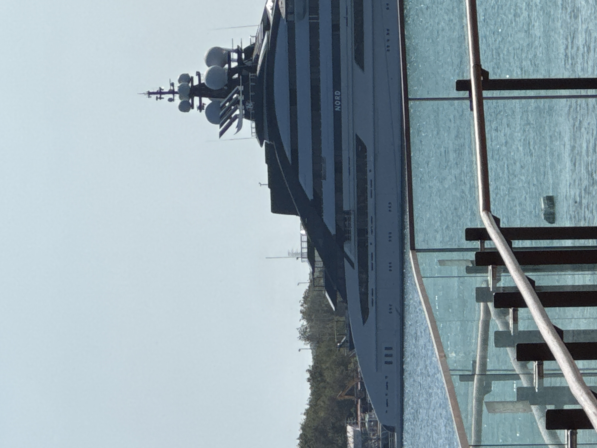 A massive, grey superyacht named "NORD" is docked in the harbor of an unknown city in Indonesia. The yacht, which features a distinctive dark, layered superstructure, is anchored in calm blue water. In the foreground, a glass railing with dark posts and light wooden handrails overlooks the harbor, suggesting a waterfront promenade or viewing area. The background is characterized by a line of green trees and a bright, clear sky. The overall scene depicts a tranquil day at a port, with the imposing superyacht as the central focus.