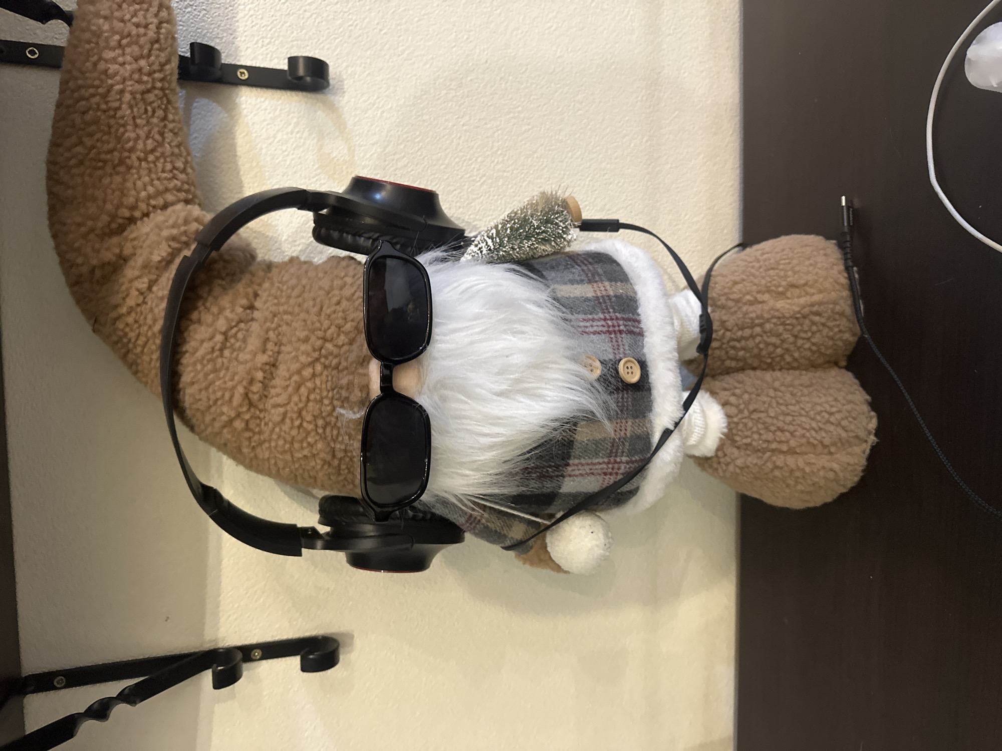The image shows a decorative gnome, likely a Christmas figurine, positioned on a dark surface against a textured, light-colored wall. The gnome is wearing black headphones and black sunglasses, and it has a long white beard and a brown fuzzy hat. Its clothing includes a plaid vest and a white trim. A small artificial Christmas tree is attached to its side, with a wire extending from it. A black audio cable is visible on the dark surface below the gnome, as well as a white cable and a white object, possibly earbuds. Two black metal brackets are mounted on the wall above and behind the gnome. The overall setting appears to be indoors, possibly a home or retail environment, with no specific geographical markers suggesting the city of Kyiv, Ukraine.