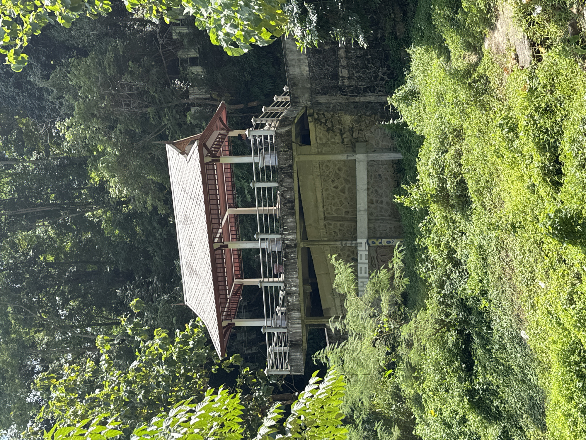 An elevated, open-air pavilion with a multi-tiered roof sits atop a concrete structure in a lush, green environment. The pavilion has a railing around its perimeter, and a person can be seen standing on it, looking out. The concrete structure is partially covered in moss and vines, suggesting it is outdoors and has been there for some time. The area around the pavilion is dense with vegetation, with trees and bushes growing thickly. The scene appears to be located in Kamala, Thailand, as indicated by the inscription "KAMALA" visible on the side of the concrete structure. The overall impression is one of a tranquil, natural setting with a man-made structure designed to offer a view of the surroundings.
