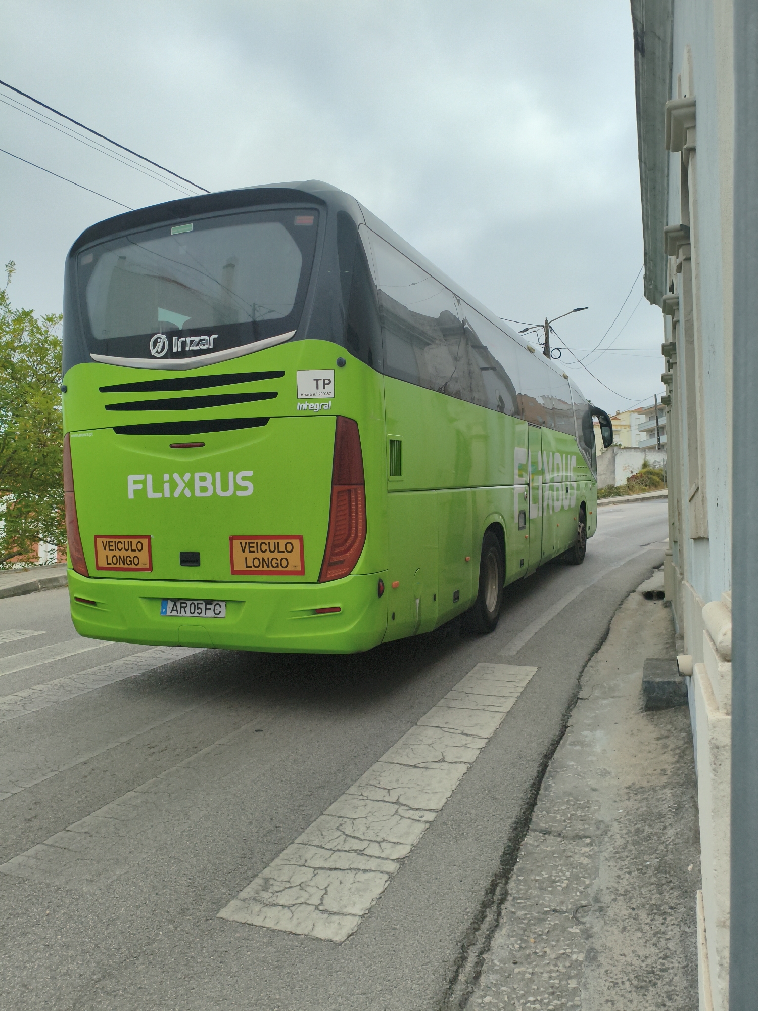 A bright green Flixbus, identified by its prominent logo and "FLIXBUS" text, is captured from a rear-side view, moving away from the camera on a street in Nazaré, Portugal. The bus features the "Irizar" logo on its upper rear. Two rectangular yellow signs with black text reading "VEICULO LONGO" are affixed to the lower rear of the bus. The license plate displays "AR05FC". The bus is on an asphalt road with a marked pedestrian crossing. To the right of the road is a weathered concrete curb and a pale blue building. Overhead, electrical wires are visible against an overcast sky. A tree with green foliage is present on the left side of the image, partially obscuring the bus. The scene is devoid of people.