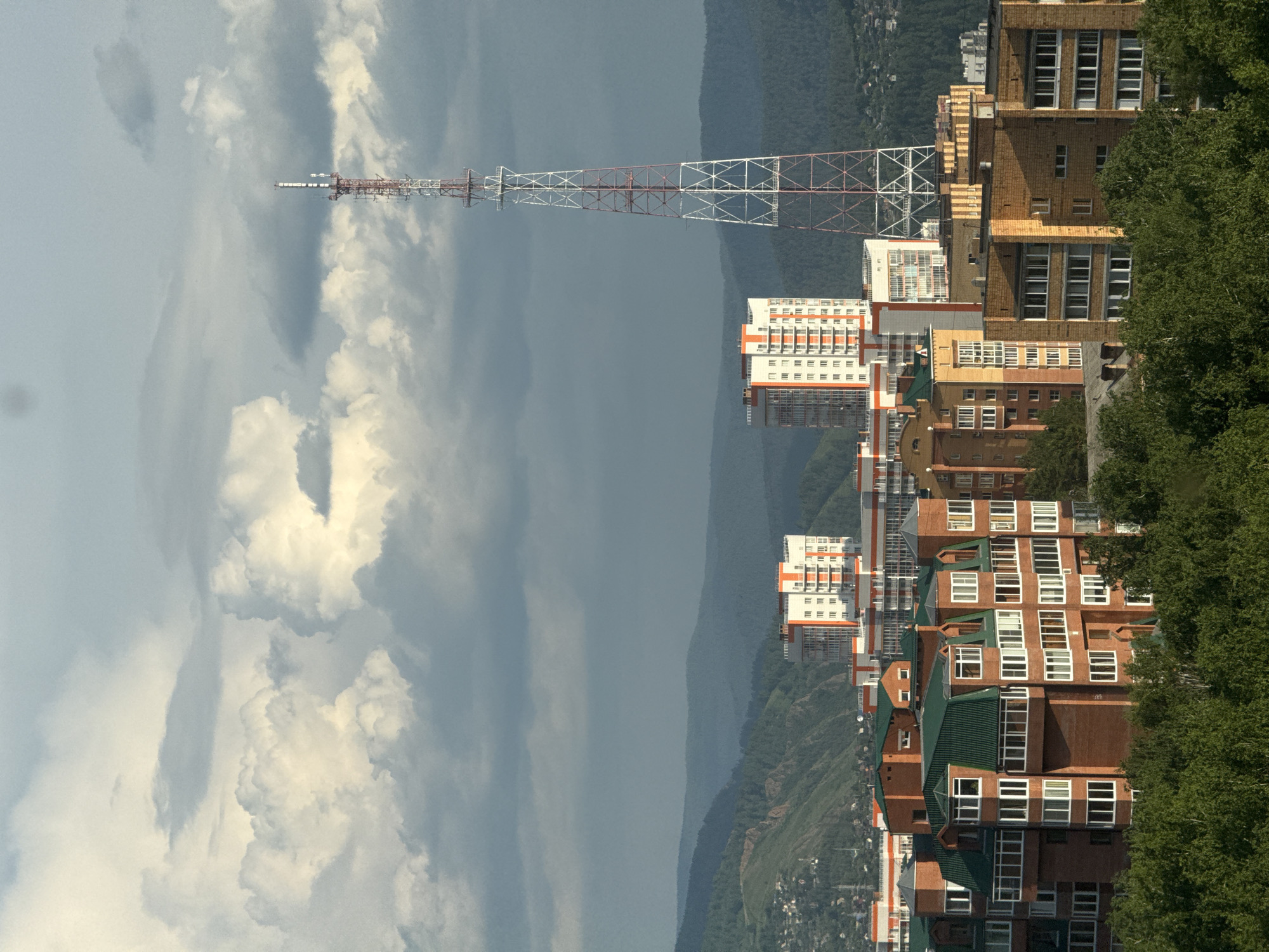 This is a medium-shot, vertical, aerial view of a cityscape in Krasnoyarsk, Russia, during daylight. The scene is dominated by a dramatic sky filled with large, white, puffy cumulus clouds interspersed with darker, gray storm clouds. In the foreground, a dense canopy of lush green trees fills the lower portion of the frame. Behind the trees, a cluster of brick apartment buildings with green roofs and numerous windows is visible. Further back, taller, modern buildings with orange and white facades stand out. A prominent, tall, lattice telecommunications tower with red and white accents rises in the mid-ground. In the background, rolling green hills, some appearing mountainous, form a layered landscape receding into the distance under the cloudy sky. The image captures a residential area with a blend of older and newer architecture, set against a natural backdrop of hills.