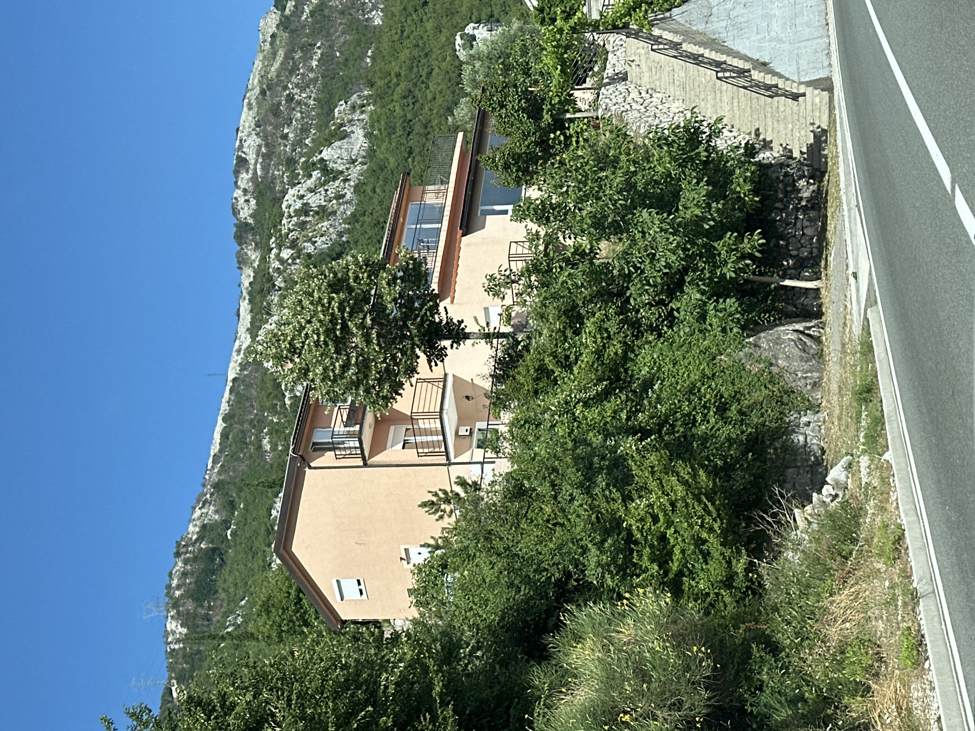 The image depicts a multi-story house situated on a hillside in Vrela, Montenegro. The house is painted a light orange and features multiple balconies with dark railings. It is surrounded by lush green vegetation, including trees and shrubs. In the foreground, a paved road with white lane markings curves to the right. To the right of the house, a stone-walled staircase ascends the hillside. The background shows a steep, rocky mountain covered in dense green trees, with the clear blue sky above. Power lines and pylons are visible on the distant mountain slope. No people are present in the image.