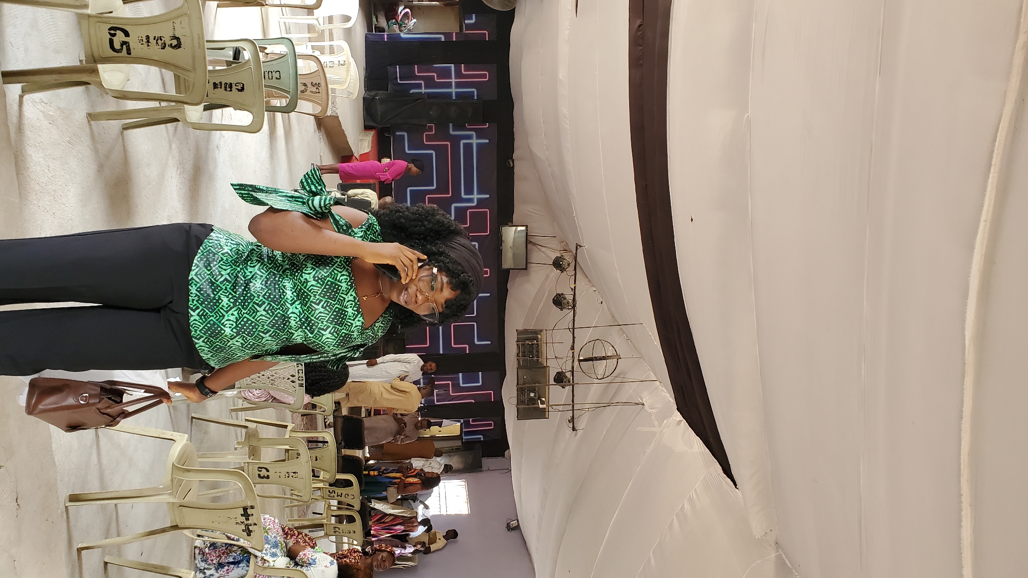 A woman in a green and black patterned top and black pants stands in the foreground of an indoor gathering, speaking on a mobile phone. She wears glasses and a black headscarf, and a brown handbag is on her right hip. Behind her, rows of light-colored plastic chairs are arranged on a concrete floor. The background features a stage with a black backdrop adorned with colorful neon geometric lines. Several other individuals are visible, some standing and others seated, suggesting a community event or service within a church or hall in Jalingo, Nigeria. The ceiling is draped with white fabric, and industrial lights and speakers are suspended above the stage.