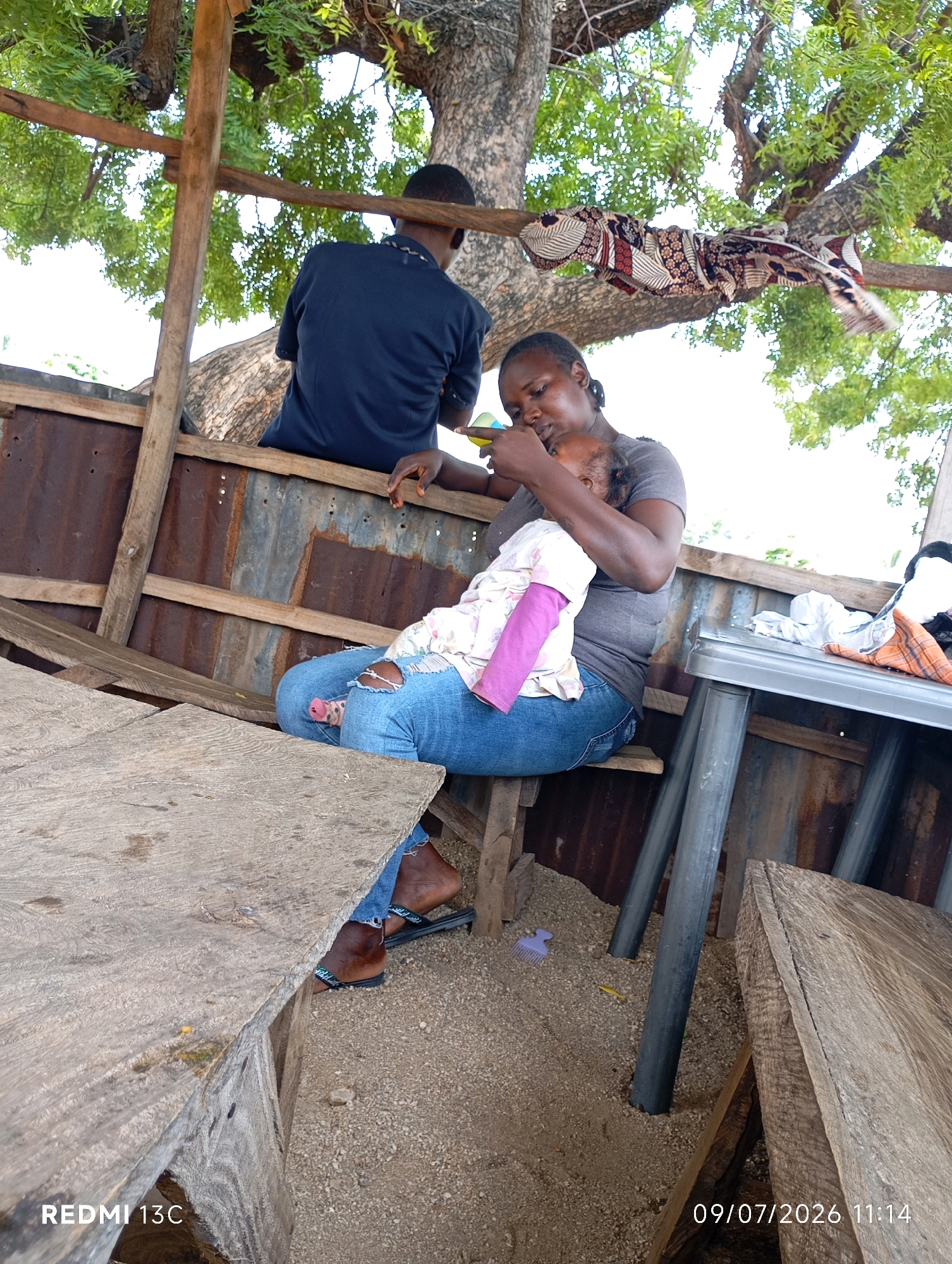 A woman is seated on a wooden bench under a tree, holding a baby and feeding it. She is wearing a grey t-shirt and blue jeans. The baby is dressed in a pink and white outfit. In the background, a person in a blue shirt is also seated, facing away from the camera. The scene appears to be an outdoor setting with a rustic shelter constructed from wood and corrugated metal. The ground is covered in gravel and sand. A wooden table is visible on the right side of the frame, with some items on it. The image is dated 09/07/2026, 11:14 and contains the text "REDMI 13C".
