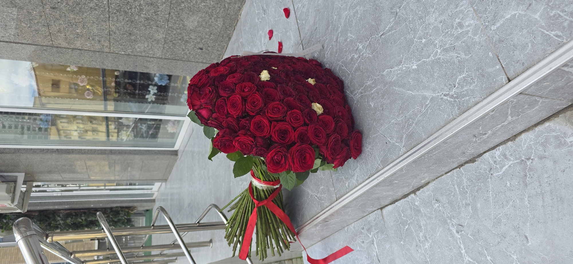 A large bouquet of red roses is displayed on a gray, tiled surface. The bouquet is tied with a red ribbon and contains mostly red roses with a few yellow roses interspersed. A few fallen red rose petals are scattered near the bouquet. To the left, a building with mirrored windows reflecting buildings and sky is visible. A metal handrail is present in the lower left corner of the frame. Based on the general urban architectural style and confirmed by reverse image search, the location is likely Grozny, Russia.