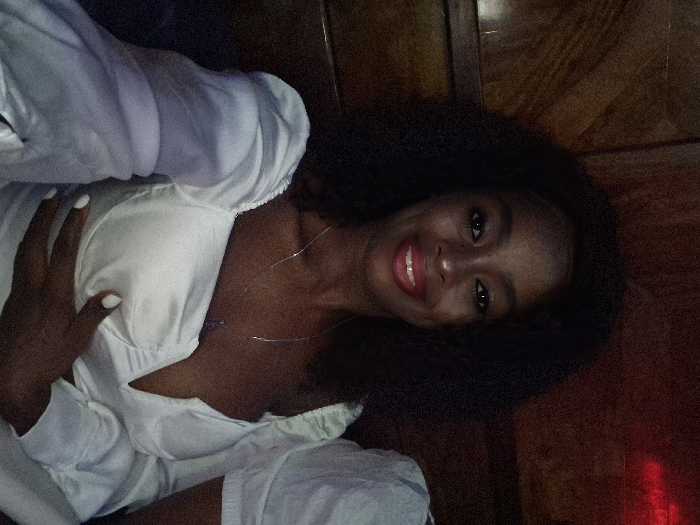 A woman with dark hair smiles warmly. She has deep brown eyes and wears red lipstick. Her nails are painted white. She is dressed in a white top, partially unbuttoned, with a silver necklace. Her right hand is casually placed on her chest. The background includes dark wooden paneling and a reddish glow on the right. The image is rotated slightly.