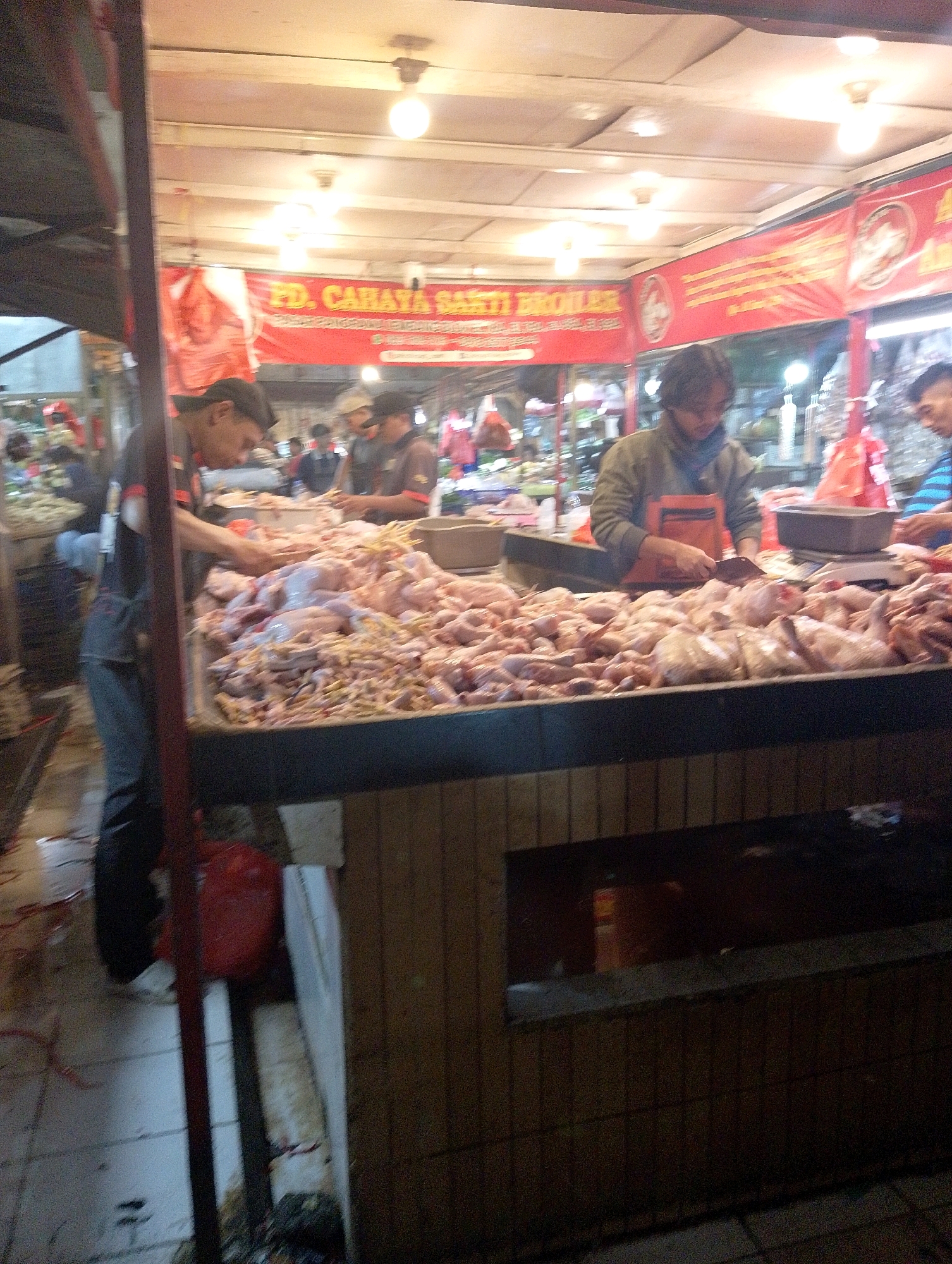 This is a medium-shot, horizontal-oriented image of a butcher stall in a market, likely in Lembang, Indonesia, given the context provided. The stall is filled with a large display of raw chickens, some whole and some cut, arranged on a counter. In the foreground, a person wearing a dark t-shirt and dark pants is seen from the waist down, with their left hand on the counter and their right hand near the chickens, appearing to be preparing them. Behind this person, a second individual, wearing a grey jacket over an orange apron and a dark scarf, is also working with the chickens, holding a knife. A red banner with yellow lettering reading "PD. CAHAYA SARTI BROILER" is prominently displayed behind the people and the chickens, indicating the name of the business. The stall has a wooden counter and shelving, and is illuminated by overhead lighting. The overall scene suggests a busy marketplace environment where fresh poultry is being sold.