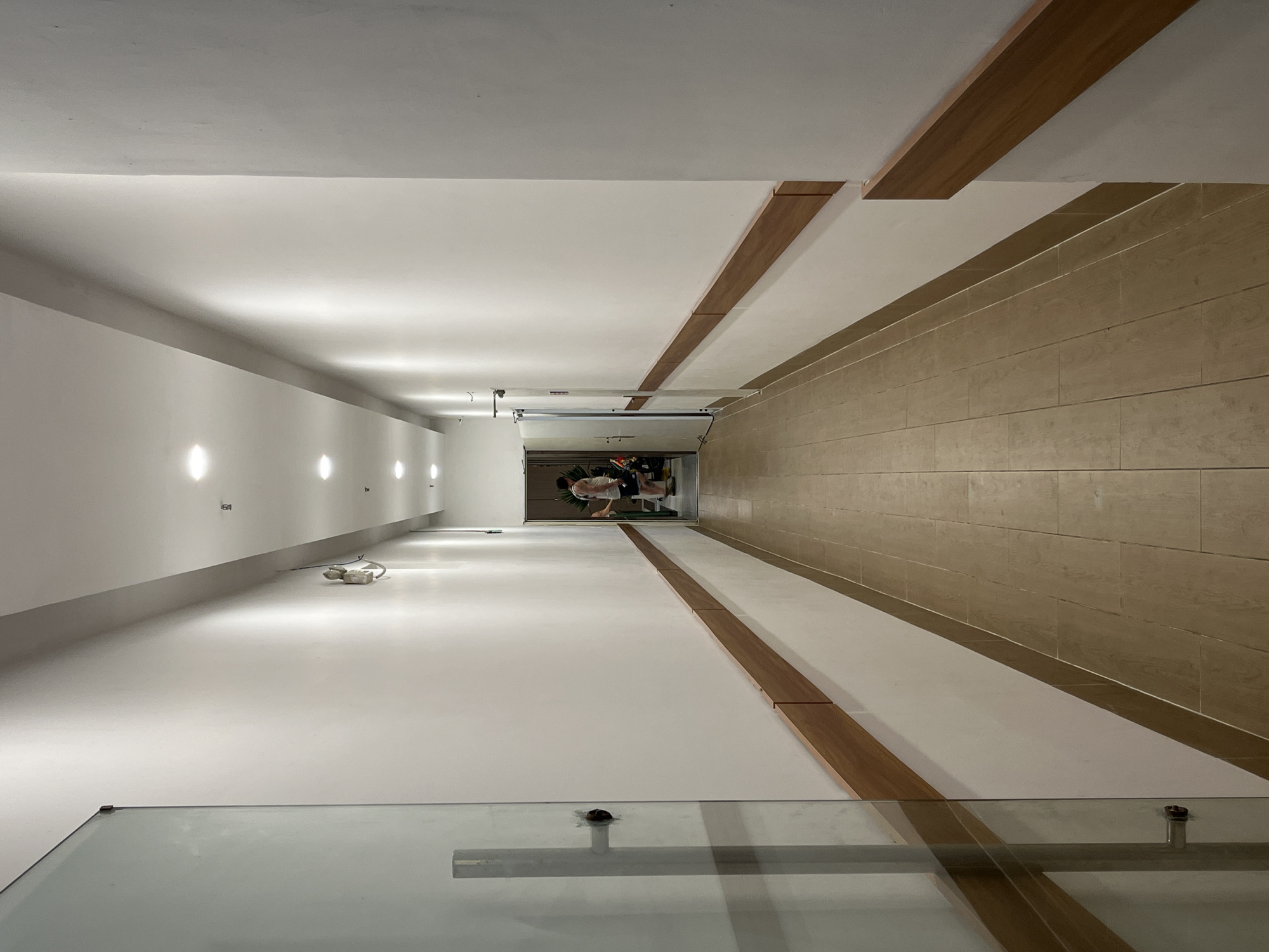 The image presents an indoor hallway with distinct design features. The left wall is white, adorned with evenly spaced sconce lights. A gray accent strip runs along the bottom of the left wall. The right wall is covered in tan-colored, horizontally oriented tiles, with a wooden accent trim between the white ceiling and tiled wall. The floor appears to be white. In the distance, at the end of the hallway, is a partially visible garage or storage area. A person is slightly stooped over, possibly organizing or retrieving items within that space. The scene is possibly a modern house, interior. The bottom of the image has a portion of a clear glass barrier, indicating a possible balcony or stairwell. Based on the prompt, the location is Bac Nha Trang Ward, Vietnam.
