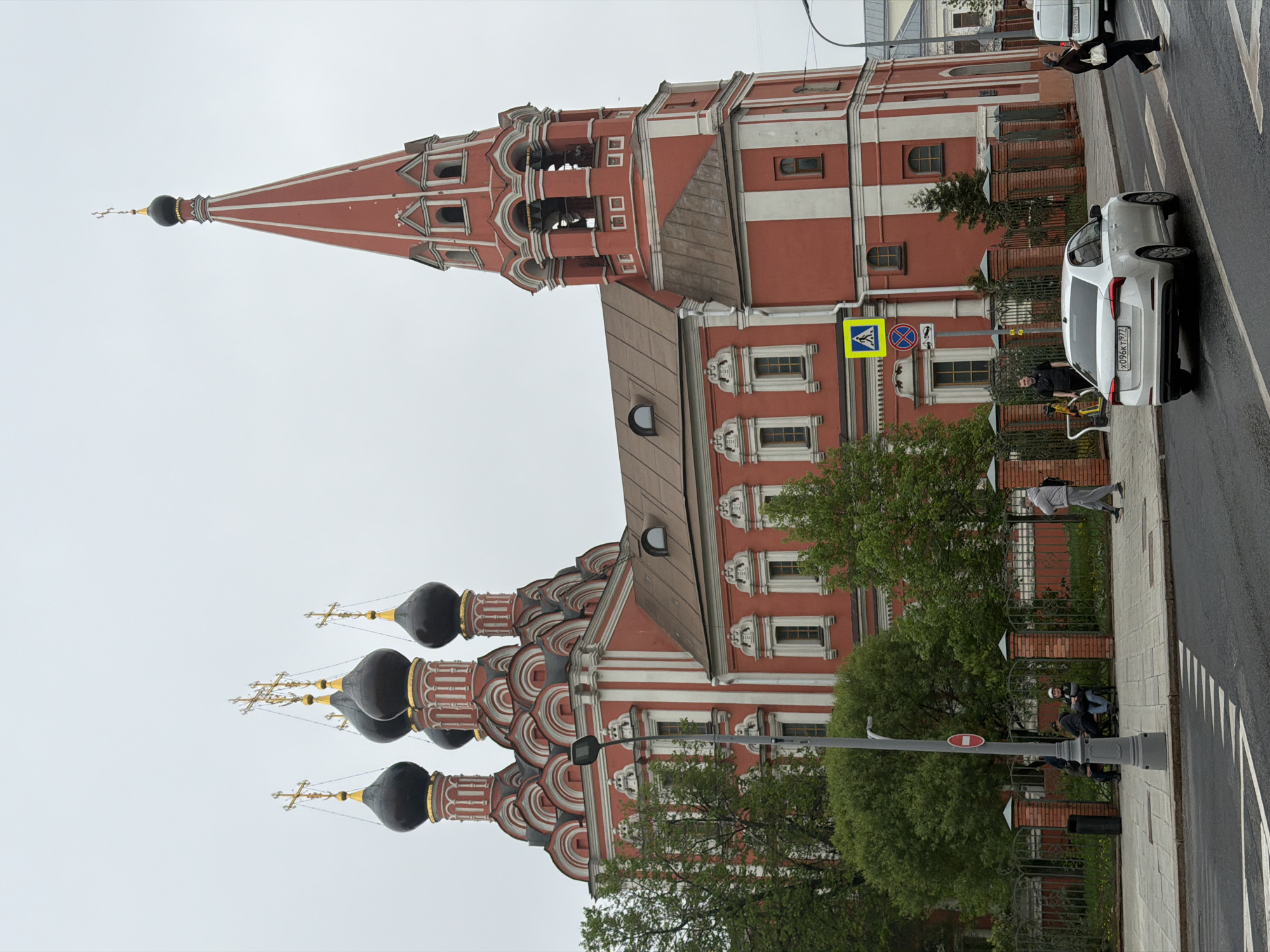 The image shows an architectural structure that appears to be a church, possibly of Russian Orthodox design, with red and white walls and multiple domes topped with golden crosses. The building has a distinctive spire. The scene is urban, as indicated by the presence of an asphalt street and a sidewalk.
A white car is parked on the side of the street. People are visible on the sidewalk, some of whom appear to be walking or interacting with the car. A street sign, identifiable by the pedestrian crossing symbol, suggests a crosswalk is nearby. Trees and other vegetation are also part of the scene. The weather seems overcast, with a uniformly grey sky.