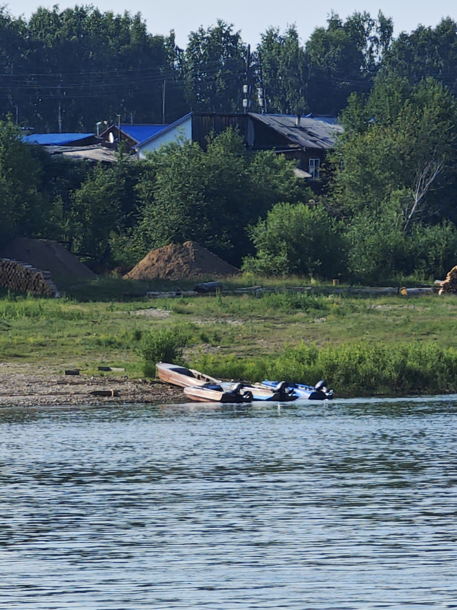The image shows a wide, calm river with a gravelly bank on the right. Several boats are moored along the bank, some partially submerged. In the background, a line of trees separates the river from a village with several houses and outbuildings. One house has a blue roof, and another has a dark, weathered roof. Piles of sand and stacked logs are visible on the bank behind the boats. The sky is clear and the overall impression is one of a quiet, rural setting. The specific location of Верхнепашино, Russia, is inferred from the context provided by the user.