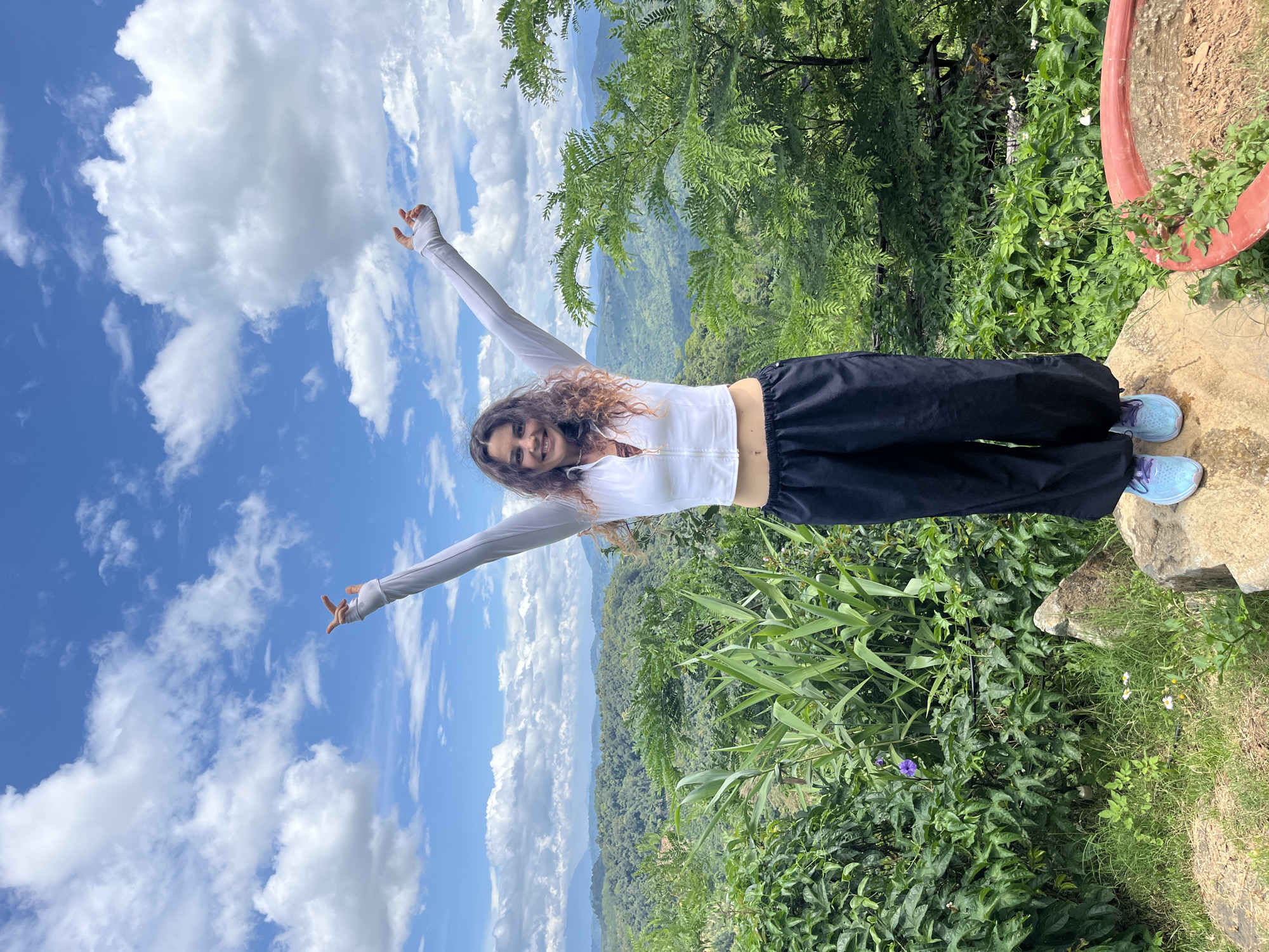 A woman stands on a rock outdoors, with her arms raised in celebration against a backdrop of a blue sky filled with white clouds and a lush green mountain landscape. She is wearing a white zip-up jacket and loose black pants, with blue sneakers on her feet. The foreground shows dense green foliage, including tall stalks of bamboo and a single purple flower, as well as some rocks. In the background, rolling hills covered in dense vegetation extend into the distance, under a bright, partly cloudy sky. The scene appears to be in a natural, elevated setting, likely offering a panoramic view of the surrounding countryside.