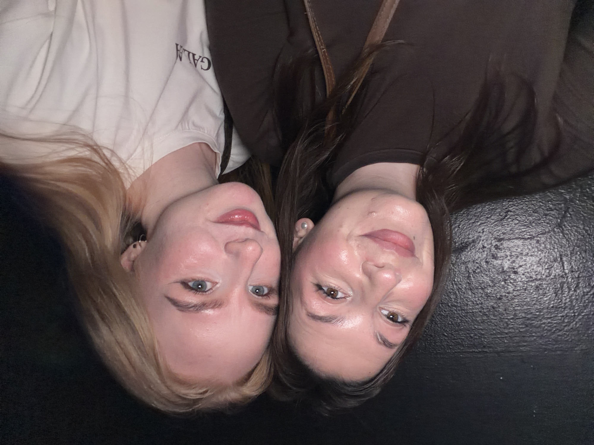 A close-up, top-down shot captures two young women lying with their heads together, looking up towards the camera with soft, content smiles. The image is rotated 90 degrees counter-clockwise, showing them positioned on a dark, textured surface.
On the left side of the frame, a woman with long, blonde hair and fair skin features light blue eyes and pink-tinted lips. She wears small hoop earrings and a light-colored (possibly white or off-white) t-shirt. The word "GALA" is clearly visible on her shirt, appearing upside down and mirrored due to the image's orientation.
On the right, another woman with long, dark brown hair and fair skin has brown eyes and similarly pink-tinted lips. She wears a dark brown top or dress, and a pearl stud earring is visible in her right ear. A strap, possibly from a bag or her clothing, is seen near her shoulder.
Both women's faces are illuminated by soft, frontal lighting, highlighting their features and makeup. Their expressions convey a sense of closeness and warmth. The background is uniformly dark, suggesting an indoor setting or a plain dark surface. The overall scene captures a friendly, casual moment between the two individuals, located in Perm, Russia.