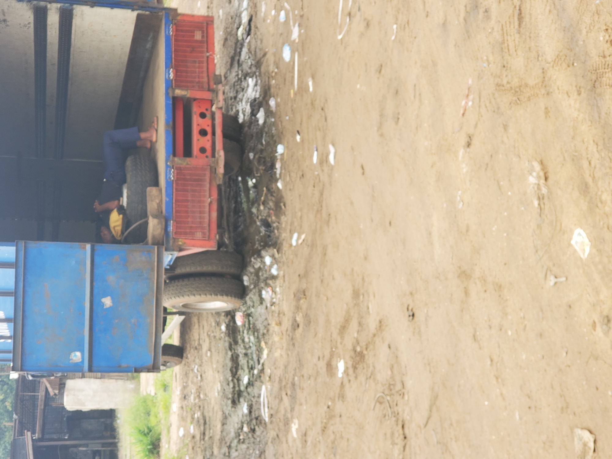 The image shows a man sleeping inside a blue and red truck, specifically within the Fegge area of Nigeria. The man is resting on a spare tire inside the truck. Outside the truck, the ground appears to be dirt and sand, and scattered with garbage. Buildings and greenery are visible in the distance.