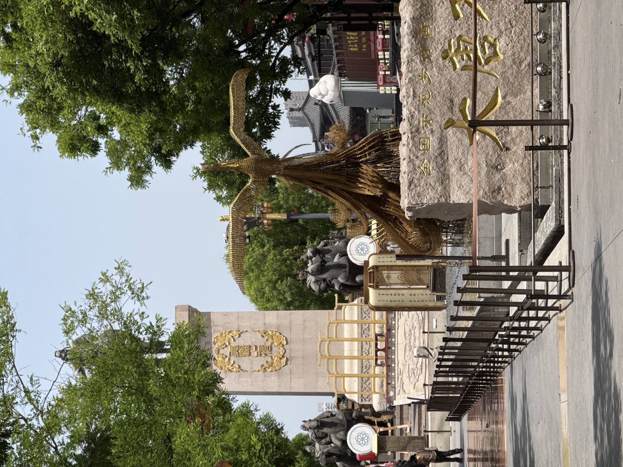 The image depicts a street scene in Yanta District, China, showcasing cultural elements and urban infrastructure. A golden phoenix sculpture stands prominently, seemingly constructed from wire or metal mesh. Behind it, a stone monument displays calligraphic characters. Trees frame the view, with glimpses of buildings and structures typical of the area. The street is paved and features metal barriers, suggesting a public space. People are visible in the lower portion of the image, moving about. The scene is captured in daylight, with a clear sky visible above the trees.