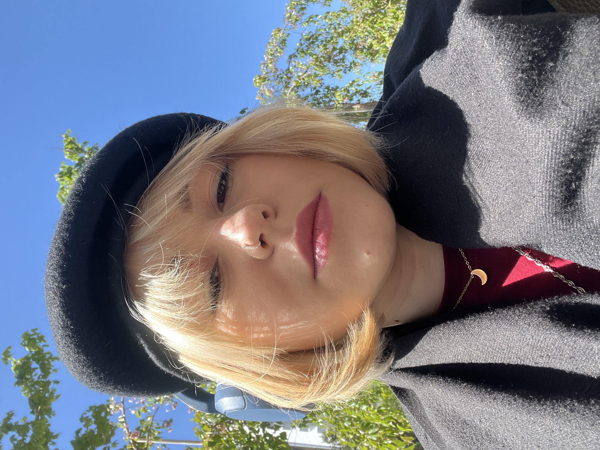 This close-up portrait, rotated 90 degrees clockwise, features a person looking directly at the camera with a neutral expression. The individual has fair skin, short, straight blonde hair, and wears pink lipstick and a small silver nose ring on their left nostril. They are dressed in a black beret, a dark red top, and a dark grey or black textured jacket or coat. A delicate gold-colored chain necklace with a crescent moon pendant is visible around their neck, and blue wireless over-ear headphones are partially seen near their shoulder.
The setting is outdoors, beneath a clear, bright blue sky with vibrant green tree foliage and some light-colored blossoms, indicating a sunny day, likely mid-day. The strong sunlight creates distinct highlights and shadows on the subject and their clothing. No specific location cues are visible within the frame, but the context indicates the scene is in Odesa, Ukraine. There is no visible text in the image.