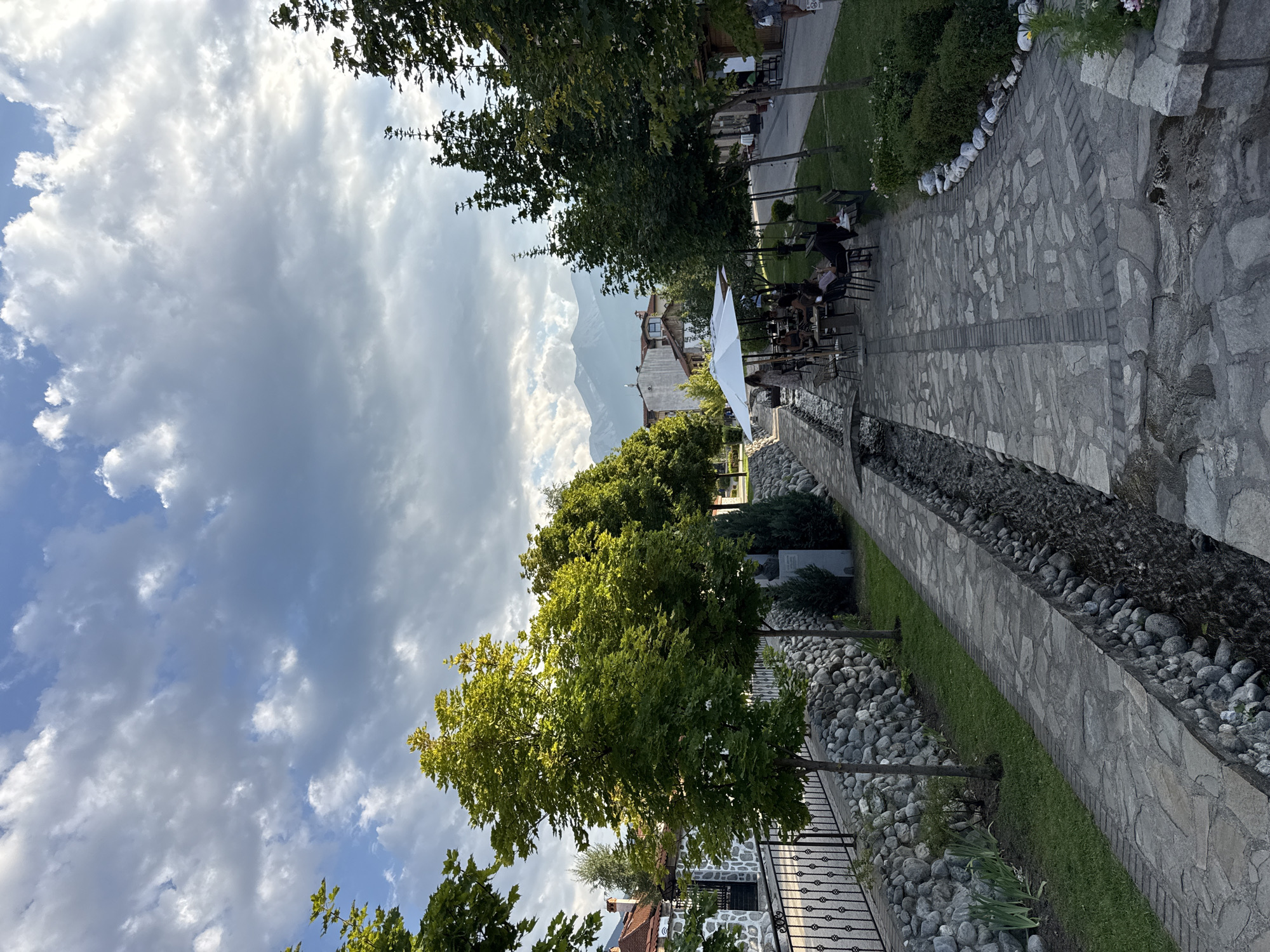 The image depicts a picturesque scene in Bansko, Bulgaria, likely along a street lined with trees and a stream. In the foreground, a stone-paved pathway follows a narrow, rocky stream. To the left of the stream, a stone wall with a black metal fence separates the pathway from a grassy area with young trees and buildings in the background. To the right of the pathway, more trees and greenery are visible, leading to an area with outdoor seating, tables, and white umbrellas, where several people are seated. Further down the street, more buildings and trees line the path, with mountains visible in the distance under a cloudy sky. One person is walking on the right side of the street.