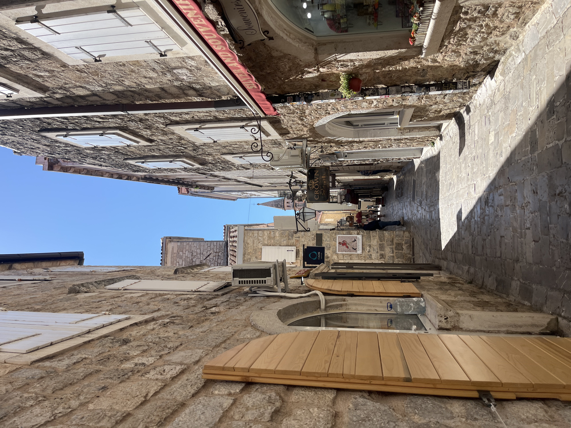 The image captures a narrow, sunlit street in the old town of Budva, Montenegro. The street is lined with buildings constructed from light-colored stone, featuring arched windows and shutters, some painted white and some natural wood. Several shops are visible, indicated by signs, including one reading "CROWN" and another for souvenirs. People are walking through the alleyway. An air conditioning unit is mounted on one of the buildings. Overhead, the sky is clear blue. The street is paved with light-colored stone, and long shadows are cast down the right side.