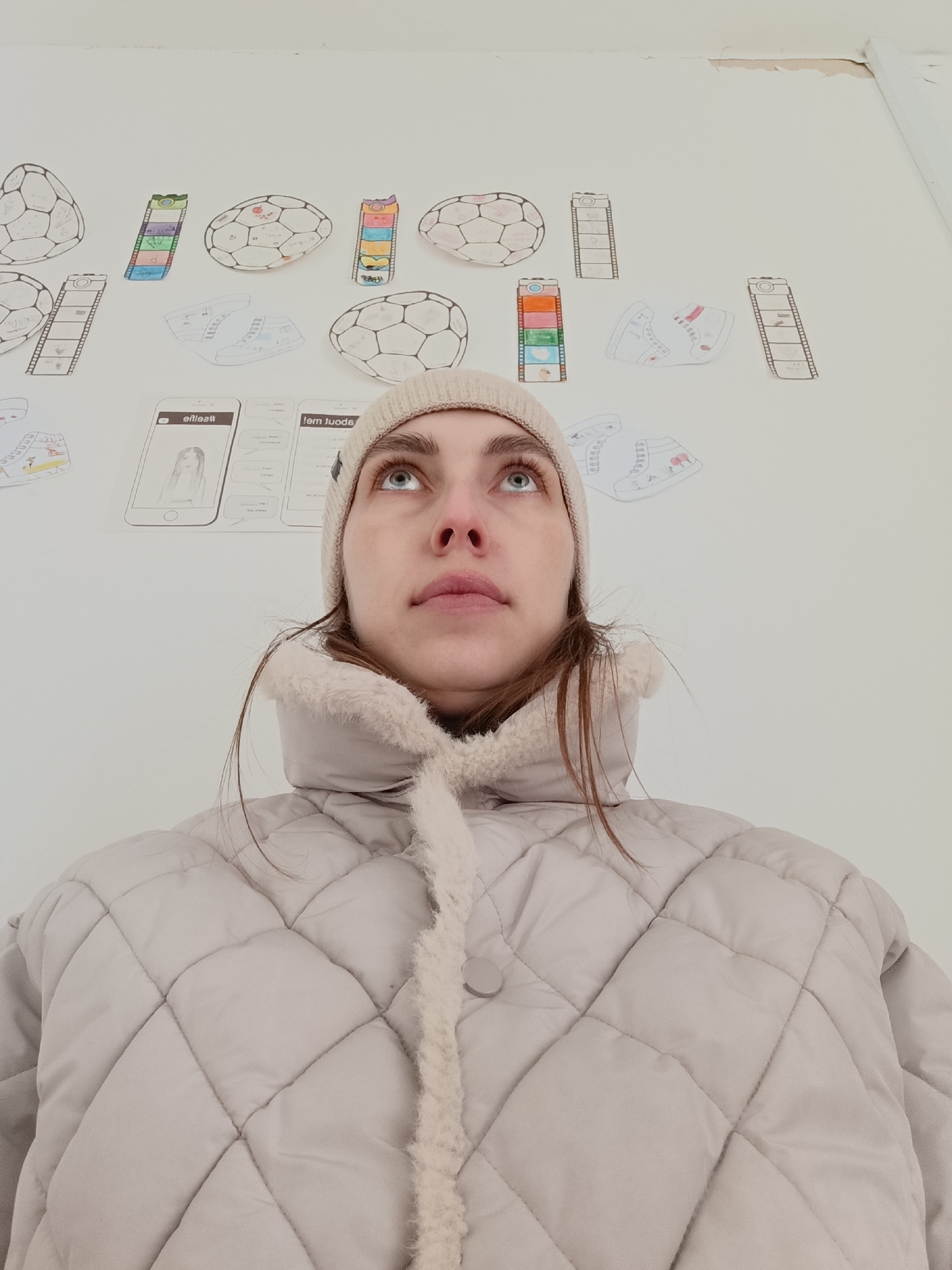 A woman with fair skin, wearing a light beige knit hat and a light beige quilted jacket with a faux fur collar, is looking upwards in an indoor setting. The background is a white wall adorned with various drawings and sketches: soccer balls, sneakers, and images resembling film strips and mobile phone screen mockups. There is evidence of minor damage to the wall near the ceiling. The location is likely in Chaikovsky, Russia.