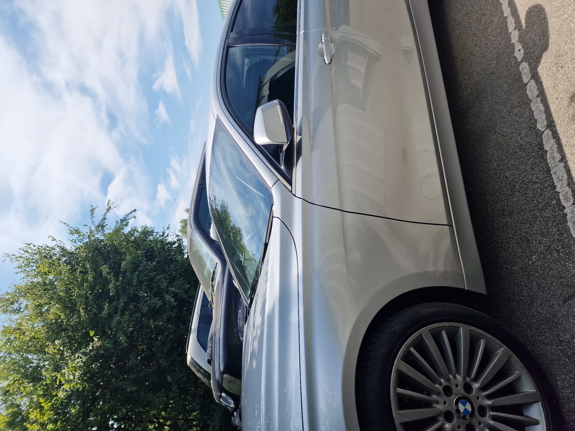 In Fenland District, United Kingdom, on a bright, sunny day with scattered clouds, a silver BMW sedan is parked on an asphalt surface. The car is seen from the side, with its front passenger door and fender in focus. The vehicle has a distinctive alloy wheel with the BMW logo prominently displayed. To the left of the BMW, partially visible, is a black SUV. Behind the cars, a dense line of lush green trees forms a natural backdrop, with the sky visible above. The scene appears to be an outdoor parking area.