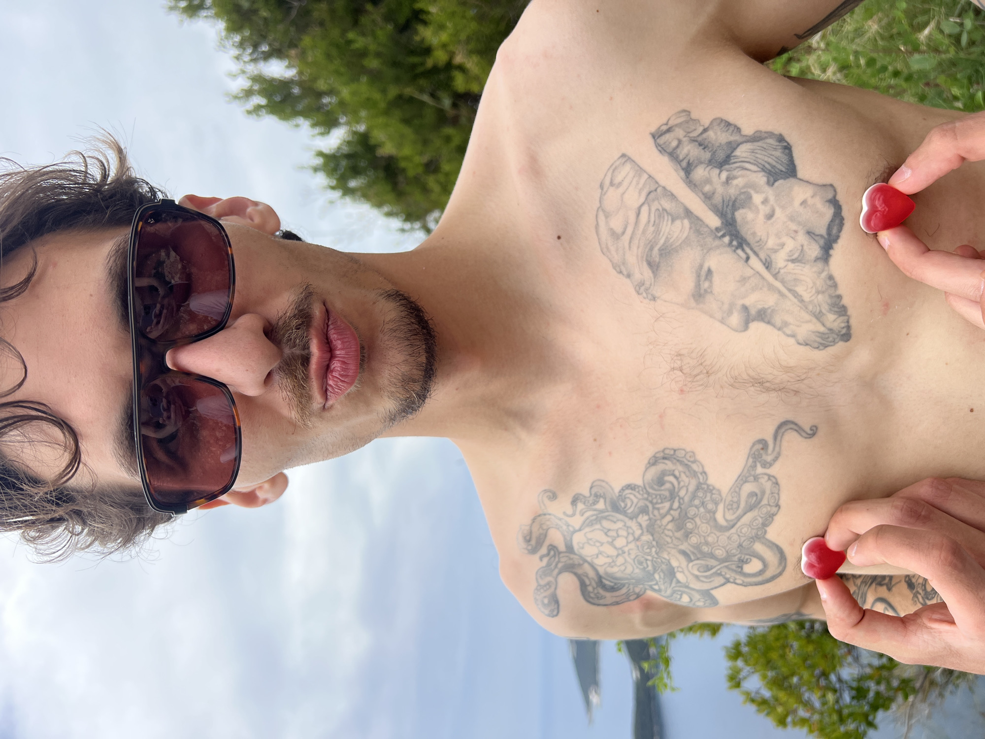 A shirtless man is holding two red heart-shaped candies against his chest. He has sunglasses on and a goatee, and he is looking directly at the camera. Two tattoos are visible on his chest: a broken bust near his right shoulder and an octopus near his left hip. Trees are in the background behind him, and the sky and water are below. The location appears to be the shoreline of Budva, Montenegro.