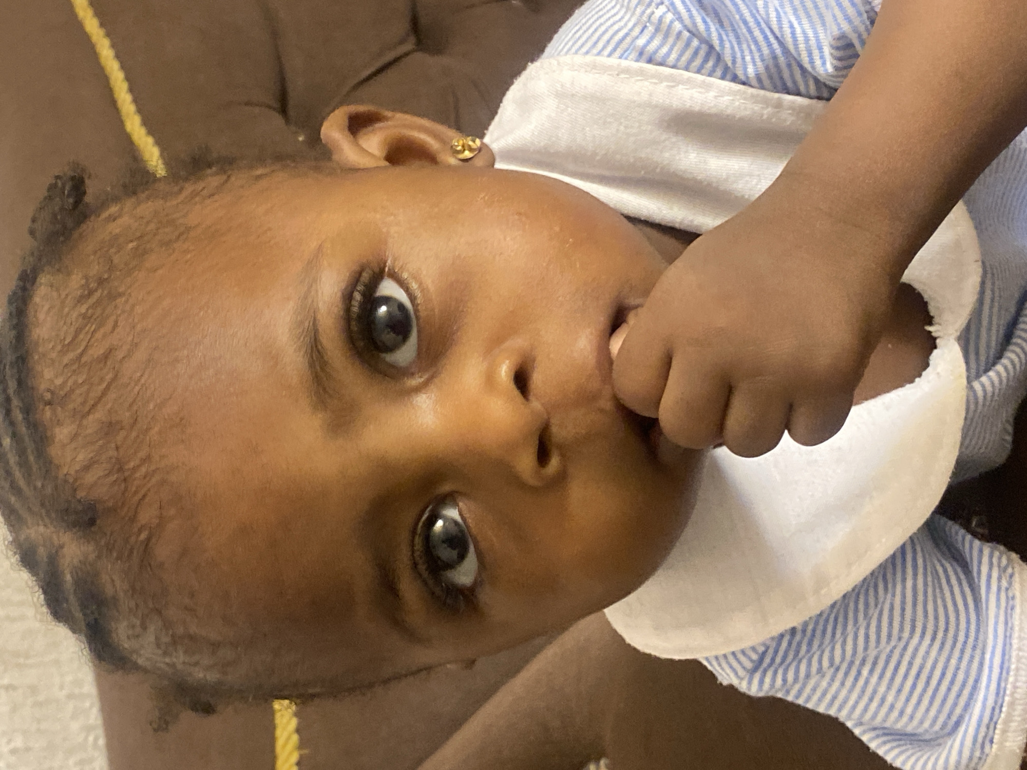 A close-up, eye-level shot shows a young Black child with braided hair and a gold earring in their right ear. The child is wearing a white bib and a blue and white striped shirt. Their right hand is near their mouth. The background is a blurred brown surface, possibly upholstery. The image appears to be taken indoors, likely in a residential setting in Abakaliki, Nigeria.