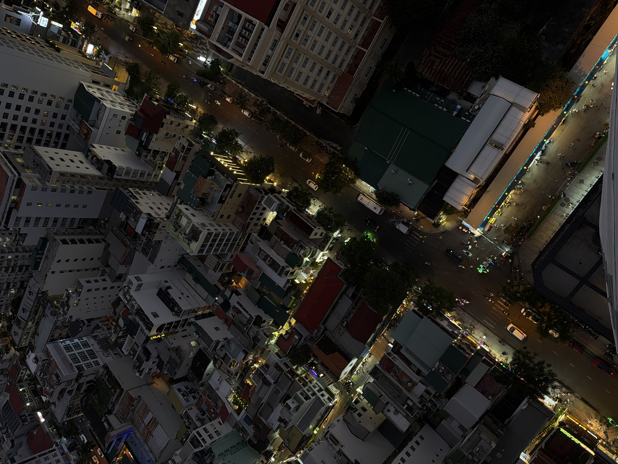 The image is an aerial night shot of Nha Trang Ward, Vietnam. The view is looking down at an angle, showcasing a dense urban landscape with buildings of varying heights and architectural styles. Streets wind between the buildings, illuminated by streetlights and vehicle headlights. Some streets are lined with trees. Pedestrians can be seen walking along brightly lit sidewalks, and cars and motorbikes move along the roadways. The overall atmosphere suggests a vibrant, active city at night.