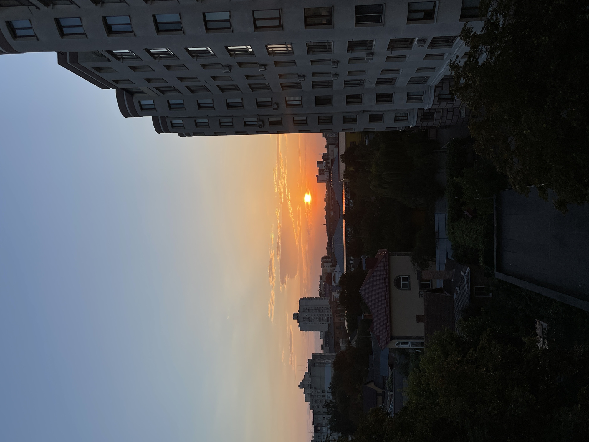 This is a photograph taken at sunset in the city of Khmelnytskyi, Ukraine. The image captures a view of buildings and trees against a colorful sky. In the foreground, on the right side of the frame, is a tall, modern apartment building with a curved facade and numerous windows. The building's architecture is characterized by rounded sections and balconies. To the left of the apartment building, the sky is a gradient of pale blue near the top, transitioning to soft orange and yellow hues closer to the horizon, indicative of a setting sun. The sun itself is a bright orange orb, low in the sky, partially obscured by wispy clouds.
Further into the scene, a cityscape unfolds with various buildings of different sizes and architectural styles, including what appear to be older residential blocks and some houses with pitched roofs. Green trees are interspersed throughout the landscape, some appearing dense and mature, partially obscuring the lower portions of the buildings. The overall impression is of an urban environment during the golden hour, with the natural beauty of the sunset contrasting with the man-made structures. There are no people or animals visible in the photograph, nor any specific actions or interactions taking place. The image is a static representation of a city view at dusk.