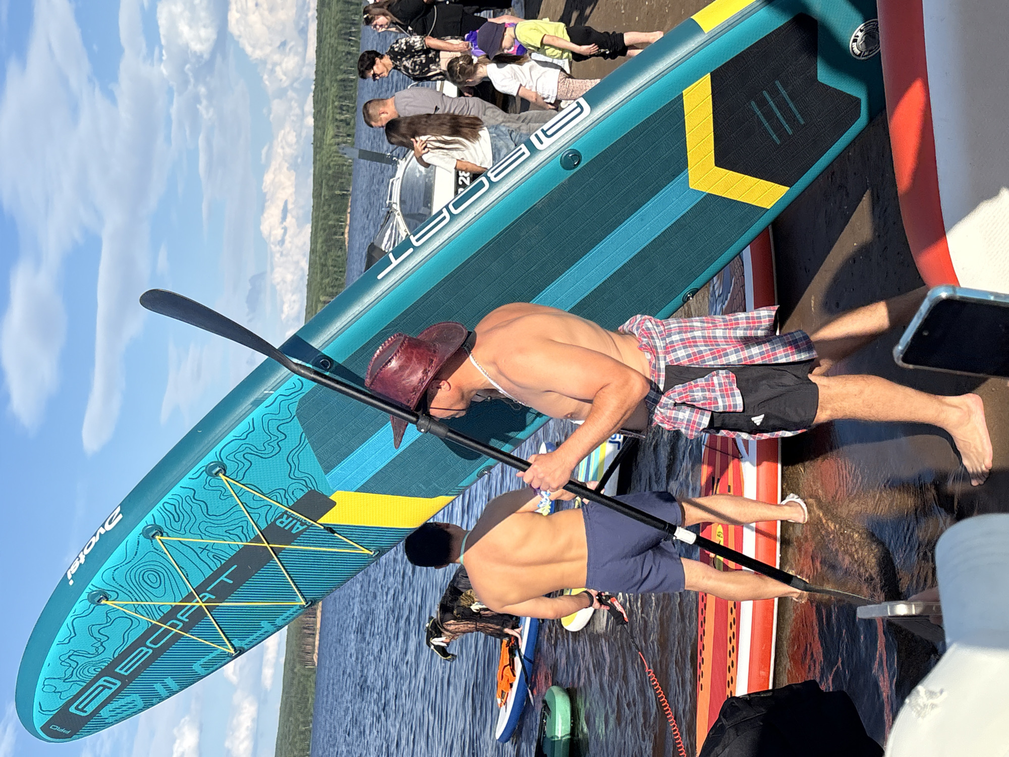 A group of people are gathered on the shore of a lake with several stand-up paddleboards (SUPs) nearby. A large turquoise and yellow SUP is propped up diagonally in the foreground, with its branding visible. Two men are holding a paddle for the SUP. One man, wearing a red hat and plaid shirt tied around his waist, is facing away from the camera. The other man, wearing blue shorts, is facing away from the camera and standing in the water. Further back, a dog wearing a life jacket is on another SUP. Other individuals, including women and children, are standing on the shore or in the shallow water. The environment is a lake with trees in the background under a partly cloudy sky. The location is the Kama River beach in Chaikovsky, Russia.