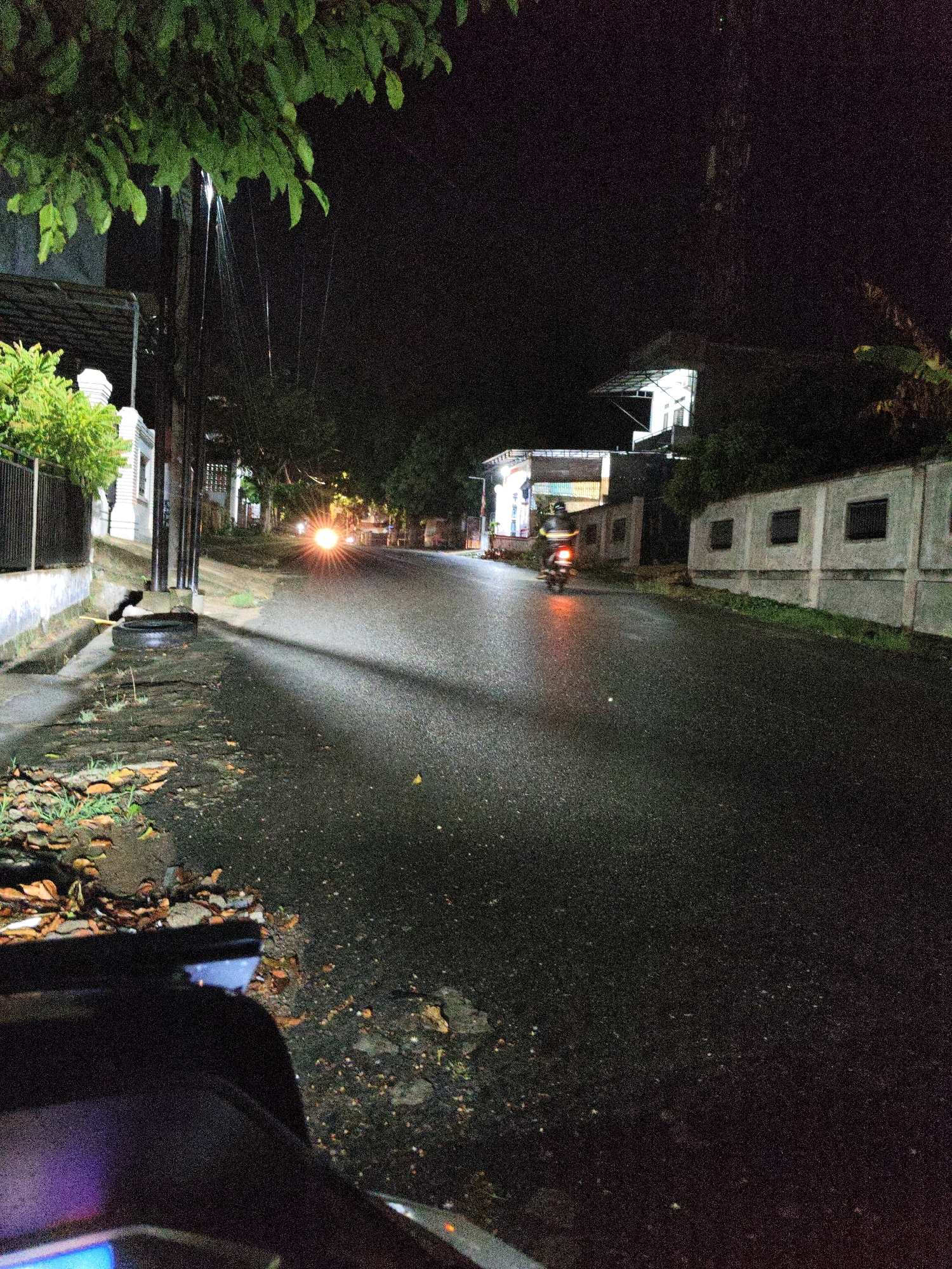 A nighttime street scene in Lhokseumawe, Indonesia, depicts a paved road with patches of wetness reflecting streetlights and vehicle headlights. On the left side of the road, a sidewalk is visible with some vegetation, fallen leaves, and a discarded tire. Across the street, buildings with rectangular windows line the side. A motorcycle is seen in motion on the road, with its headlights illuminated. The overall ambiance suggests a residential or commercial area at night, possibly after rain.