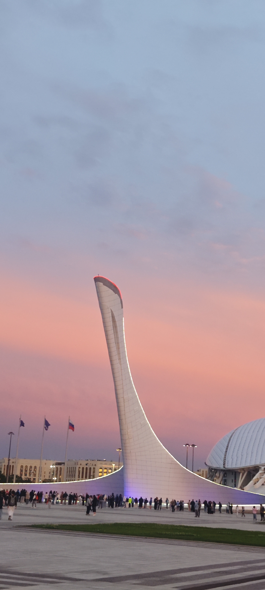 The image shows the Sochi Olympic Cauldron in Sirius, Russia at dusk. The sky has a mix of pink and blue hues. The cauldron is a tall, white structure with a red top. A crowd of people stands around the base of the cauldron. There are flags and buildings in the background.