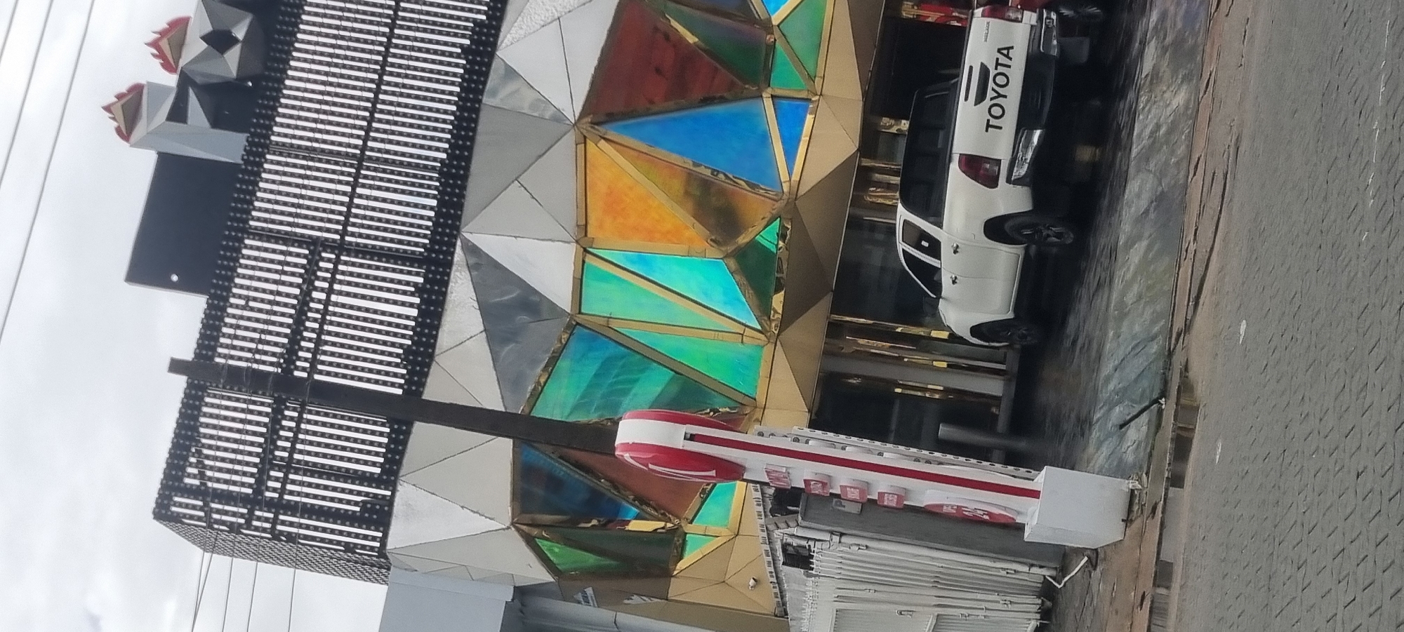 This is a rotated, eye-level, medium shot of a modern building facade and a white Toyota pickup truck parked on a paved surface. The building features a geometric design with triangular panels in various colors, including iridescent blues, greens, and oranges, as well as metallic silver and gray. To the left, a dark, perforated structure extends upwards, with some decorative red and gray elements at the top. In front of the building, a red and white structure, possibly a sign or display, is visible. The Toyota pickup truck, with "TOYOTA" clearly visible on its tailgate, is positioned to the right of the building, partially obscured by the colorful facade. The ground appears wet, suggesting recent rain or a damp environment. The sky, visible in the upper left portion of the image, is overcast, indicating a cloudy day. The scene appears to be in Moba, Nigeria, and there is no discernible activity or emotion depicted. The overall impression is a contemporary urban landscape with striking architectural elements and a vehicle.