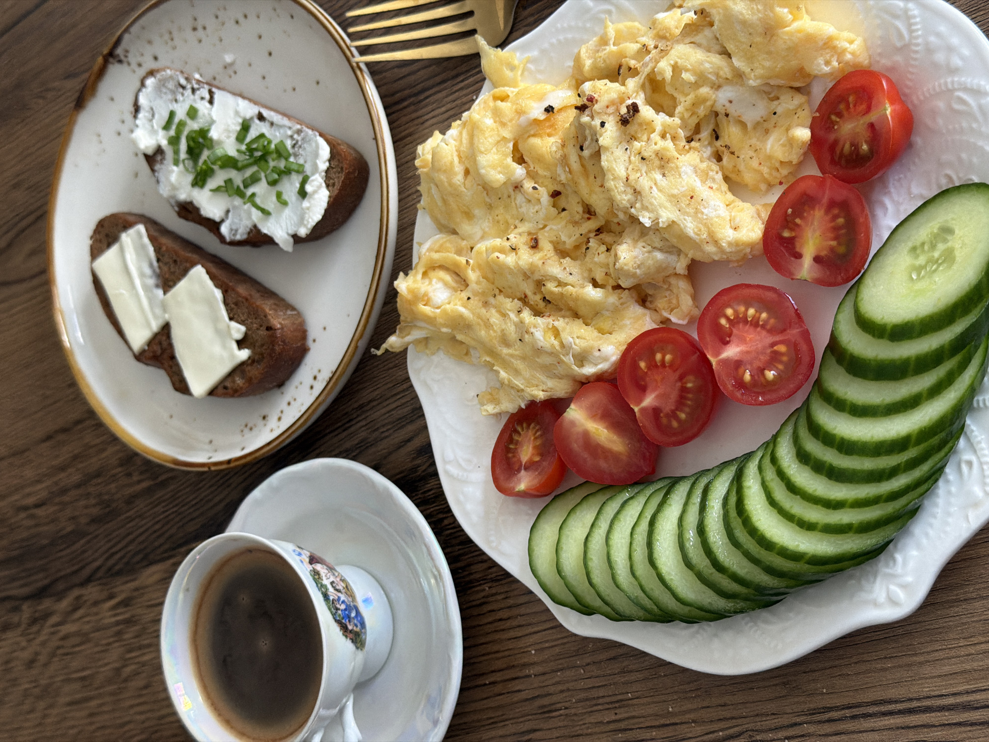 A breakfast scene is captured in an overhead shot on a wooden surface. A white plate with scalloped edges holds scrambled eggs, sliced cherry tomatoes, and thinly sliced cucumbers. Another plate, also white, contains two slices of dark bread, one topped with cream cheese and chopped green onions, the other with butter. A cup of dark coffee sits on a saucer, both adorned with a floral design. A gold-colored fork is partially visible in the upper portion of the frame. The location cannot be determined from the image.