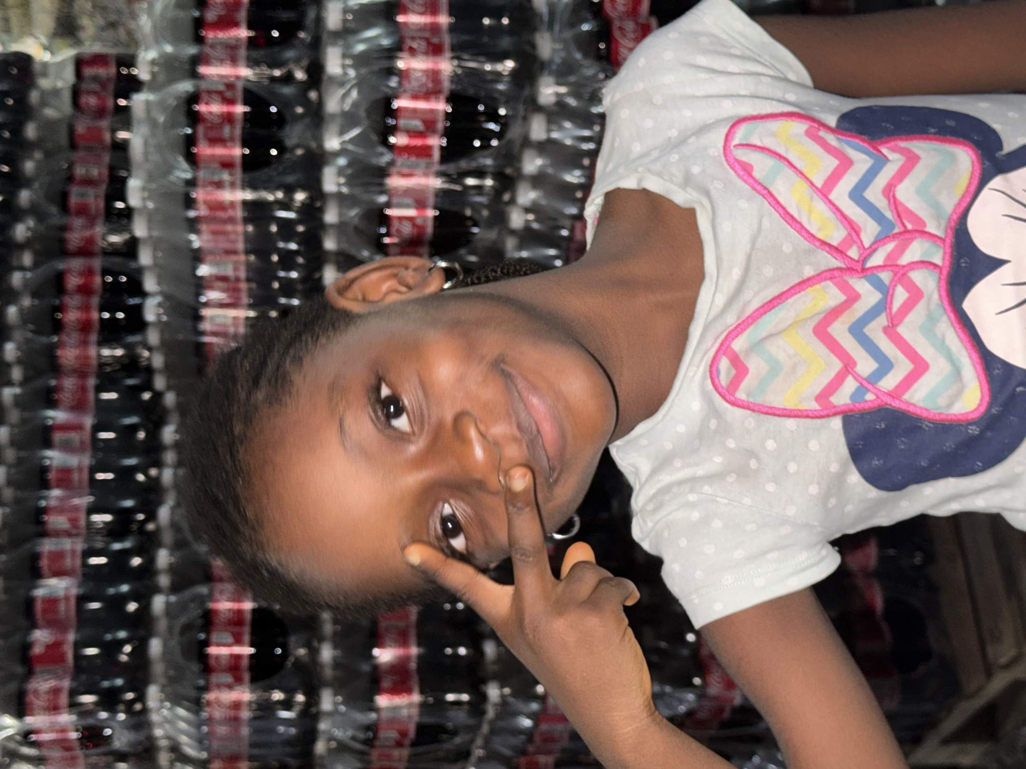 A young Black girl, approximately 5-10 years old, is shown from the chest up, smiling directly at the camera. She has short, dark hair and wears small silver hoop earrings. Her left hand is raised to her face, forming a peace sign (V-sign) with her index and middle fingers extended. She is wearing a light-colored, short-sleeved t-shirt adorned with small white polka dots and a large graphic of a bow featuring a chevron pattern in pink, yellow, and blue, outlined in bright pink. The background is dominated by a wall of stacked Coca-Cola soft drink bottles, identifiable by their dark liquid, plastic bottles, and red labels, arranged in uniform rows. A portion of a wooden pallet is visible in the lower-left corner of the frame. The scene is located in the city of Ado, Nigeria.