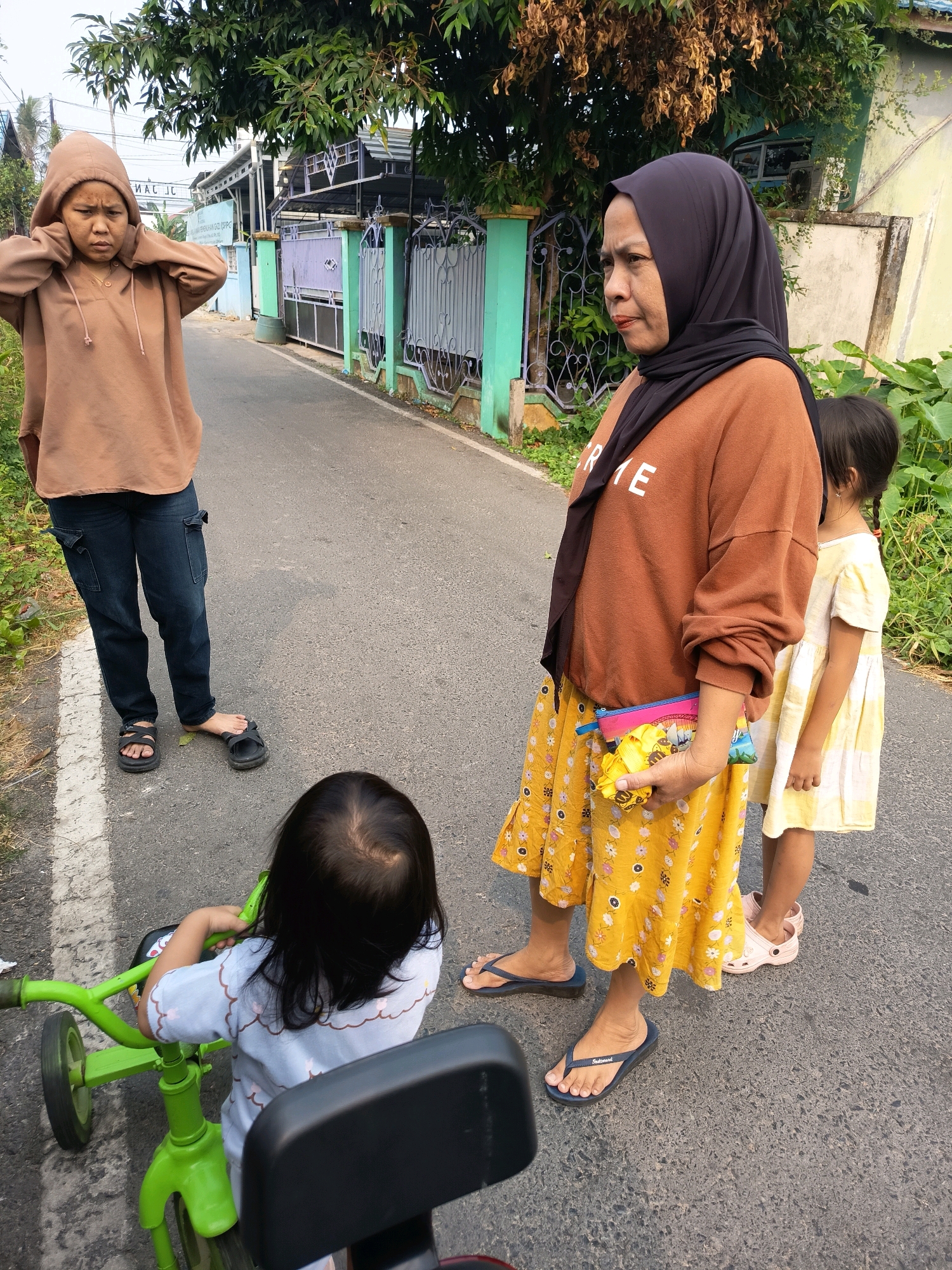 In Banjarmasin, Indonesia, a woman wearing a brown hoodie stands on a paved road, her hands on her ears. To her right, another woman in a black hijab and a brown sweatshirt looks away, holding a colorful clutch and yellow fabric. Beside her stands a young girl in a yellow dress. In the foreground, a child sits on a green tricycle. The scene is set against a backdrop of houses and lush greenery, typical of residential areas in the city.