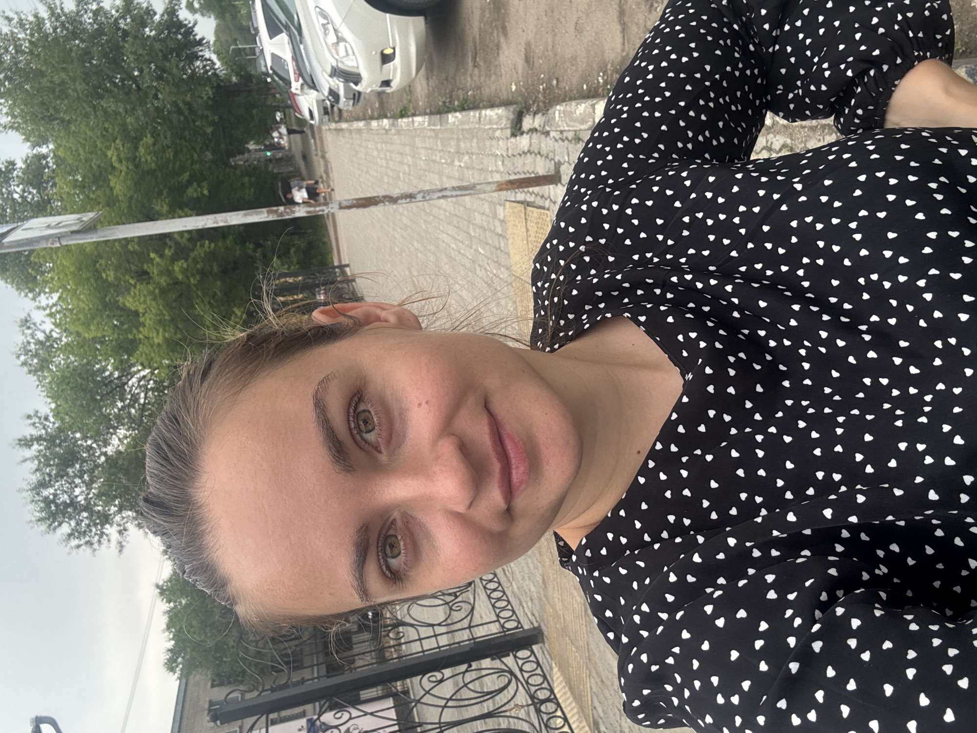 A woman with light skin, brown hair pulled back, and green eyes is taking a selfie outdoors. She is wearing a black dress with a white heart pattern. Behind her, a black metal fence with ornate scrollwork is visible, along with a sidewalk and parked cars. Trees and a building can be seen in the background. The sky is overcast. The presence of a traffic light and the general urban streetscape suggest this is likely a street in the city of Blagoveshchensk, Russia.