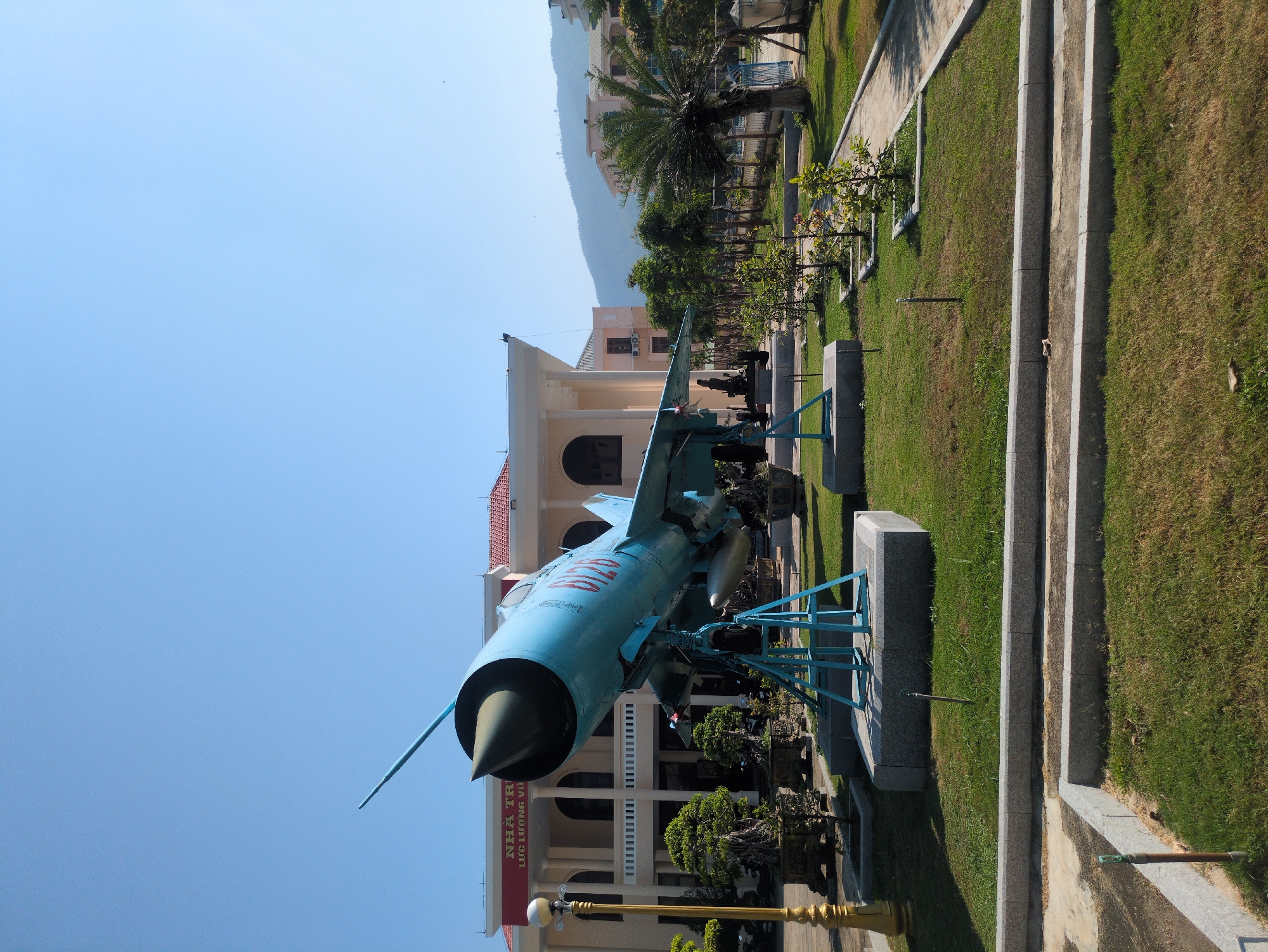 The image shows a blue MiG-21 fighter jet mounted on a blue metal display stand in a park-like setting. The jet is positioned in front of a white building with arched windows and a red roof. A red banner with white lettering hangs on the building. Green grass and trees surround the display. In the background are buildings and a mountain. The location is South Quy Nhon Ward, Vietnam.