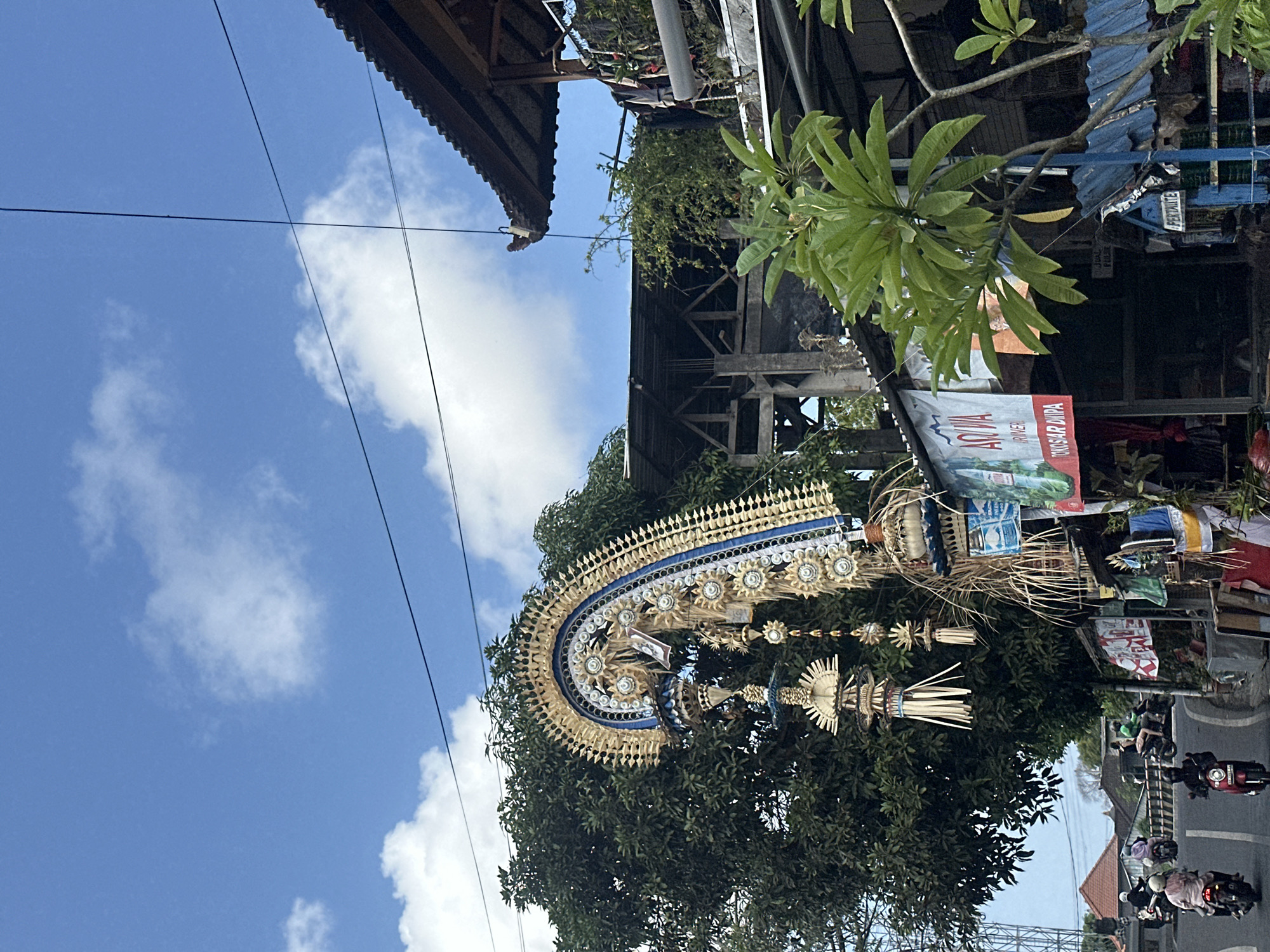 This image captures a street scene in Denpasar, Indonesia, likely near a residential or commercial area given the presence of shops and houses. A large, ornate traditional Balinese decoration, possibly a Penjor or a similar festive structure, dominates the left side of the frame, arching over the road. It's adorned with straw, woven materials, and blue and white accents. A lush green tree provides a backdrop to this decoration.
Below the decoration and the tree, a small shop or warung is visible, with various wares displayed, including signs for "Aqua" water and "Toko Sar Dipa." A blue structure, potentially a small fuel kiosk with "Pertamina" branding, is also present. In the background, more buildings and vegetation are seen.
The road in the foreground is asphalted and shows some traffic. Several motorbikes are on the road. One motorbike in the foreground is red, with a rider wearing a black helmet and jacket. Another motorbike to the left has a rider in a white helmet and pink shirt. Other motorbikes and riders are further down the road, suggesting ongoing street activity.
The environment is illuminated by daylight, with a bright blue sky filled with scattered white clouds. Electrical wires are visible crisscrossing the sky. The overall impression is of a vibrant, active street in a tropical setting.