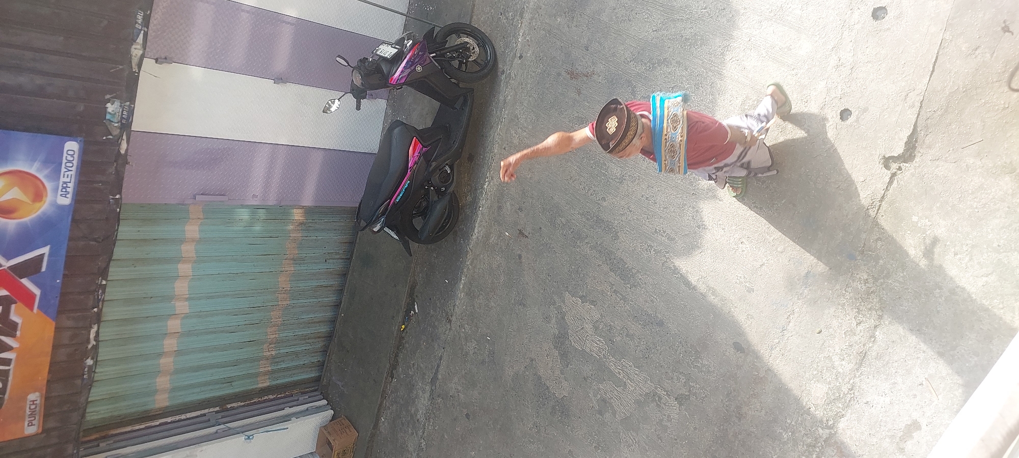 In Jayapura, Indonesia, a person wearing a red shirt, a white and patterned sarong, a black and gold patterned peci (cap), and green sandals walks on a concrete surface. A blue and gold patterned cloth is draped around their neck. Their right arm is raised, with the hand open. A black scooter with pink and purple graphics is parked to the right of the person, on an incline. In the background, a corrugated metal door painted teal with light yellow stripes is visible, along with a purple and white striped wall. A faded advertisement with the text "PUNCH" and "APPLEYOGO" is mounted above the teal door. A small cardboard box is on the ground to the left of the door. The scene is illuminated by sunlight, casting a shadow of the person on the concrete.
