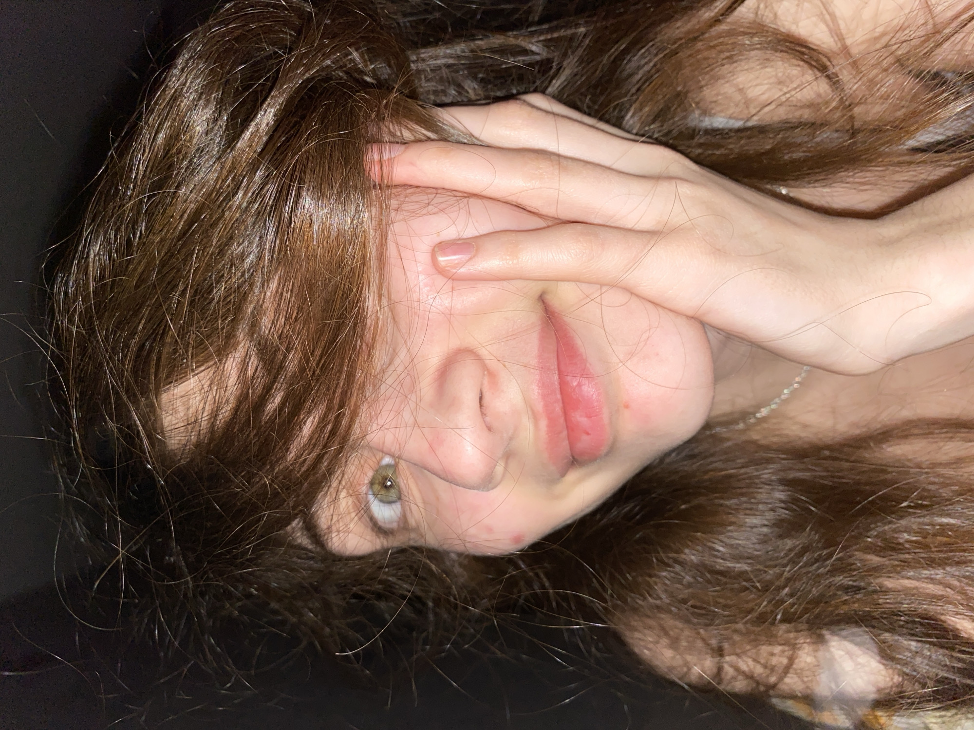 A close-up, vertically oriented photograph captures a person with long, brown hair, lying down or reclining. The subject's head is tilted to the side, with the dark, possibly shadowed background providing minimal environmental context. One hand is partially covering the face, extending across the temple and cheek, with fingers lightly touching the skin. The visible hand displays natural nails.
The person's left eye, bright green in color, is clearly visible and gazes directly at the viewer. Part of the nose and the lips, which are full and have a natural pink hue, are also discernible. The hair is spread out around the head, appearing slightly disheveled with some lighter strands catching the light. A delicate, thin silver-colored necklace chain is visible around the neck. The skin appears fair, with a few minor blemishes present on the cheek and nose area. No text is visible in the image. The overall impression is intimate, possibly suggesting a moment of rest, shyness, or a casual, candid pose.