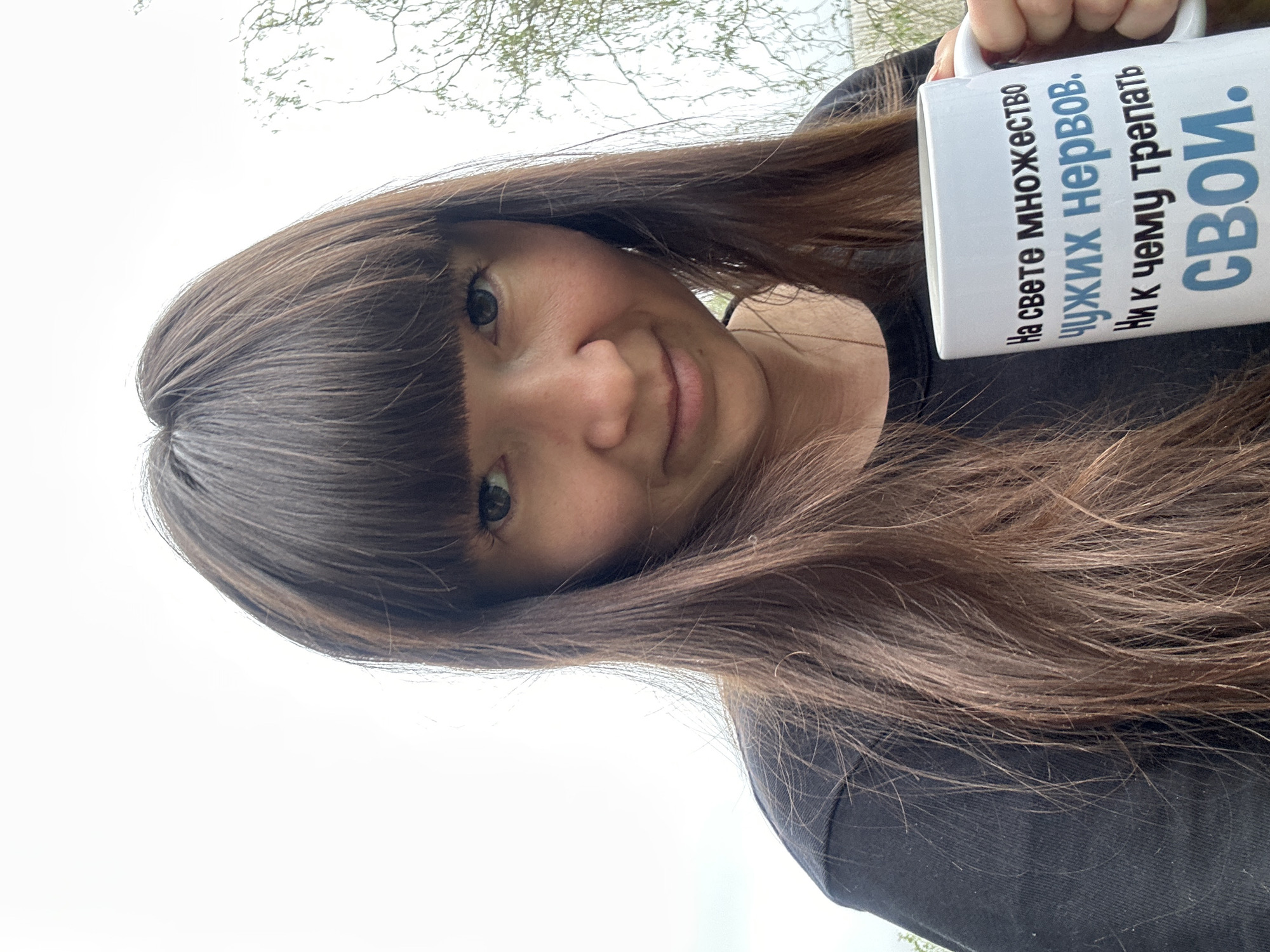 A young woman with long brown hair and bangs is holding a white mug with text in Russian. The woman is wearing a black shirt, and the background appears to be outdoors, with trees and a cloudy sky visible. The photo was taken in the city of Среднее Кечево, Russia.