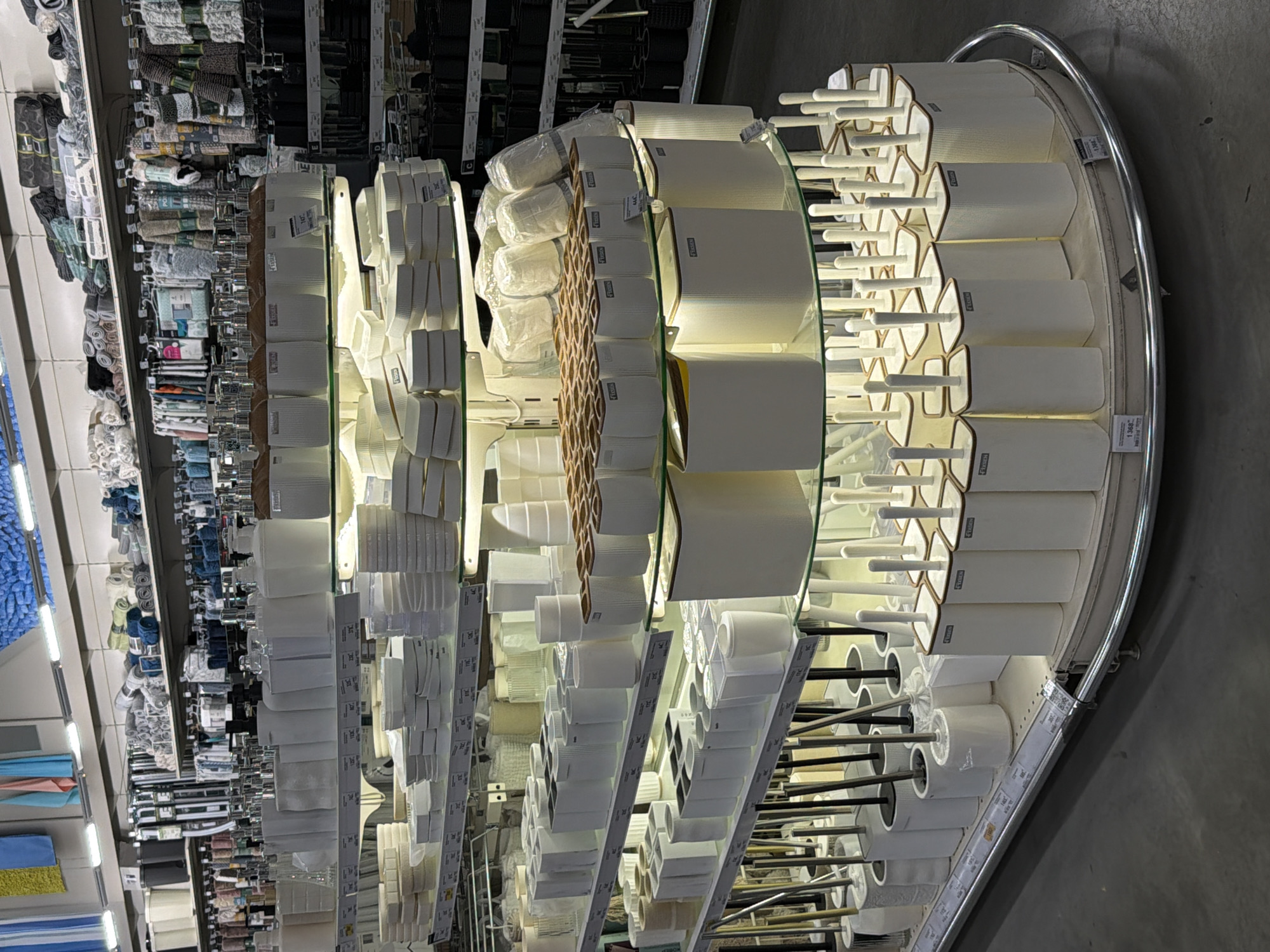 The image shows the interior of a retail store, likely in Izhevsk, Russia (based on the provided location). The focus is on a semi-circular display shelving unit filled with various bathroom accessories, predominantly white in color. These include what appear to be toilet brushes with holders, soap dispensers, and other bathroom fixtures. The shelves are made of glass, supported by metal frames. Price tags are visible on the shelves near the products. In the background, other shelves are stocked with a variety of goods, including textiles and cleaning supplies. The lighting is bright, illuminating the products on display. The floor is gray. There are no people visible in the immediate frame.