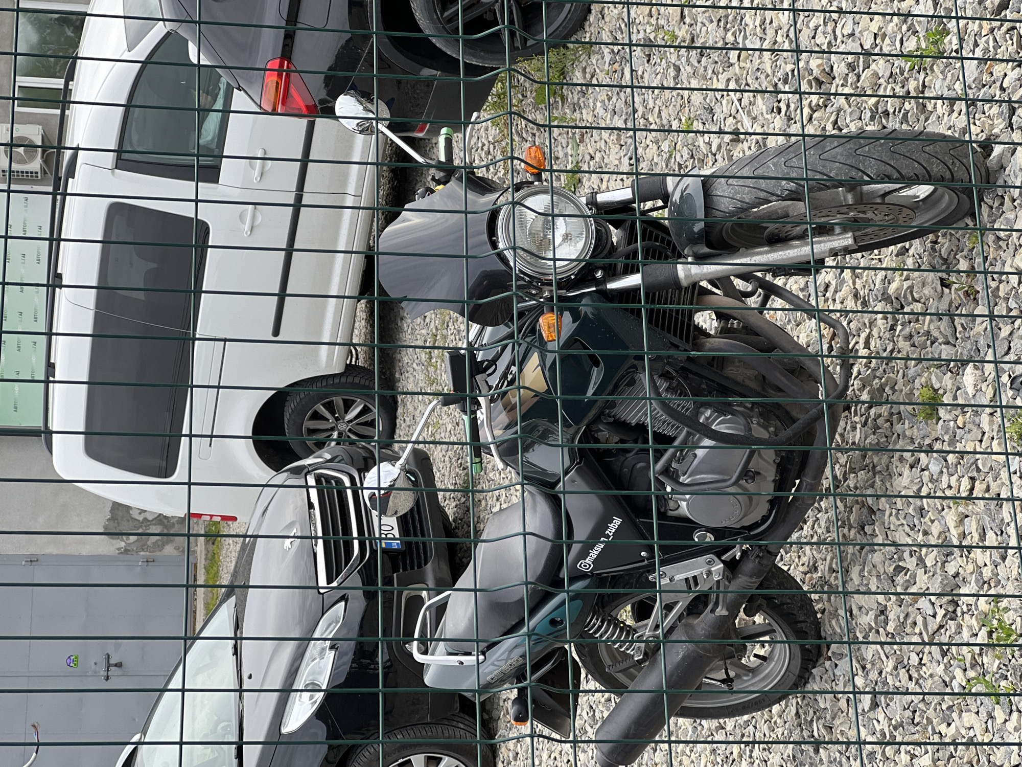 The image presents a scene in Velyki Hai, Ukraine, focusing on a motorcycle positioned behind a green wire fence. The motorcycle is a dark-colored Honda CB500, as indicated by the "CB500" marking and the engine design visible through the fence. The ground is covered in gravel, and behind the motorcycle are two parked vehicles: a white van and a dark-colored car. The vehicles and motorcycle are partially obscured by the fence. The lighting suggests it is daytime.