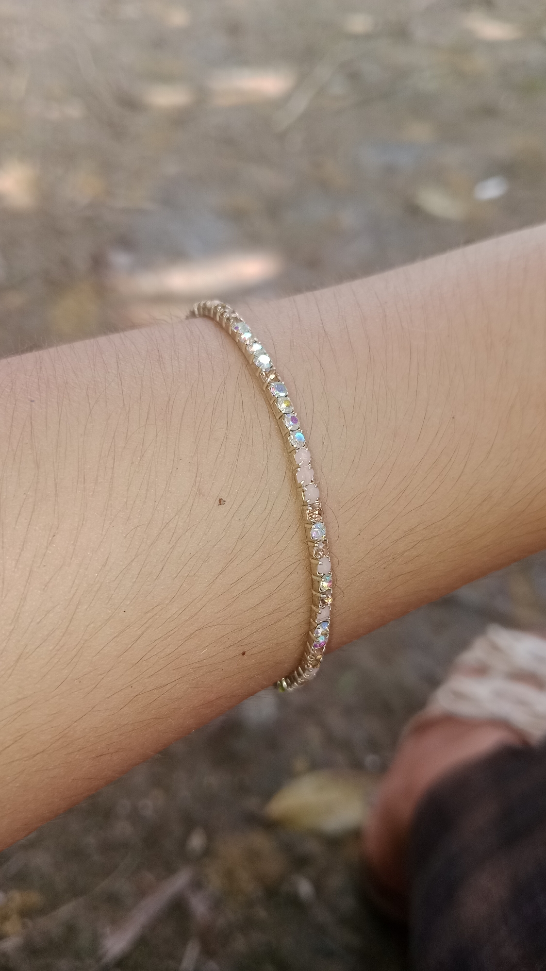 A close-up, low-angle shot captures a person's arm wearing a delicate, thin gold bracelet adorned with alternating clear and pale pink gemstones. The bracelet is positioned mid-forearm, curving gently with the limb. The arm is covered in light, fine hairs and shows some small imperfections, possibly moles. The background is blurred, suggesting an outdoor setting with muted earth tones, indicating the presence of dry foliage and possibly some ground cover. The lighting appears natural and somewhat diffused, casting soft shadows. There are no visible landmarks or contextual clues to indicate the specific location within Wono Sari, Indonesia.