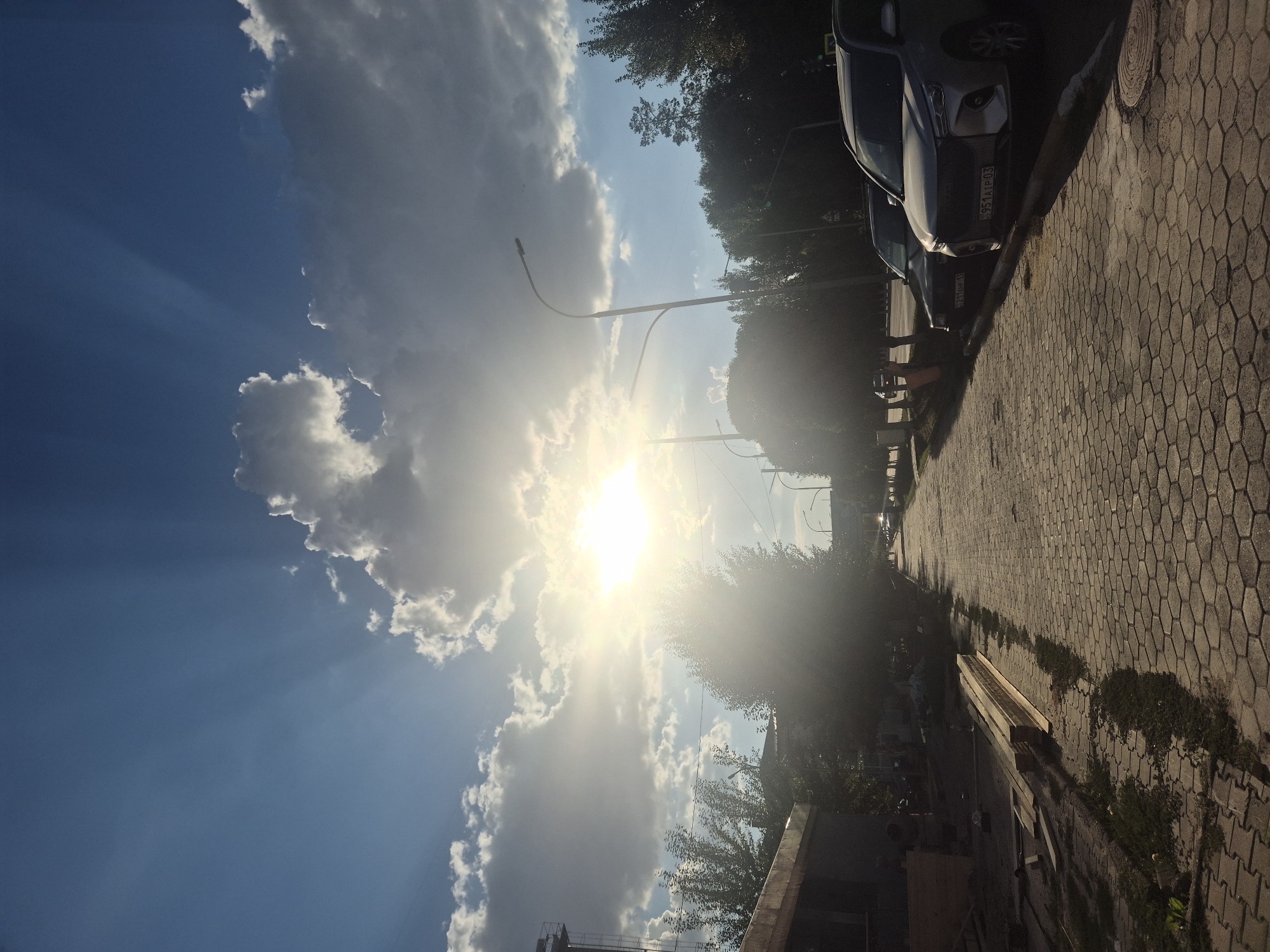 The image depicts a street scene in Kokshetau, Kazakhstan, under a bright, sunny sky with dramatic cloud formations and sunbeams. The sun is shining brightly, partially obscured by clouds, creating a lens flare effect in the center of the image.
The street is paved with hexagonal interlocking stones. On the left side of the street, there are construction materials, including stacks of lumber and what appears to be building supplies. A partially constructed building or structure is visible. On the right side of the street, cars are parked along the curb. One car in the foreground is a silver SUV, and behind it is a darker sedan.
Trees line the right side of the street, with their foliage appearing green. Streetlights are also visible along the road. The overall atmosphere suggests a daytime scene, likely late afternoon or early evening due to the angle of the sun and the quality of light. There are no people clearly visible in the frame, though some indistinct figures might be present near the construction site. The environment appears to be a typical urban street with elements of ongoing construction.